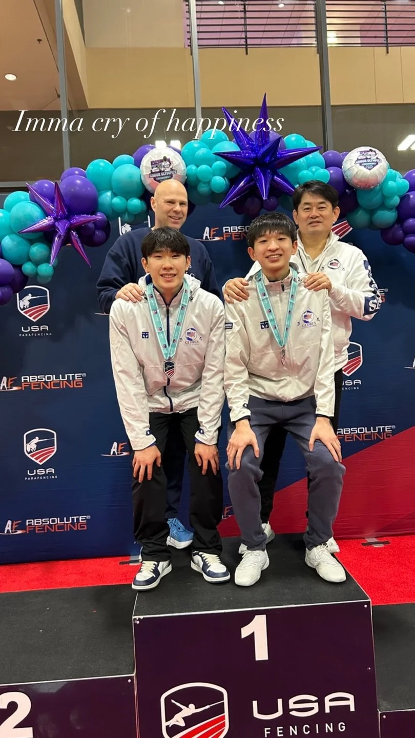 Two young male fencers in white jackets with medals around their necks, standing on a winners' podium with two adult men, all smiling. Behind them is a blue backdrop with logos and decorations, including purple and teal balloons, large purple star-sh