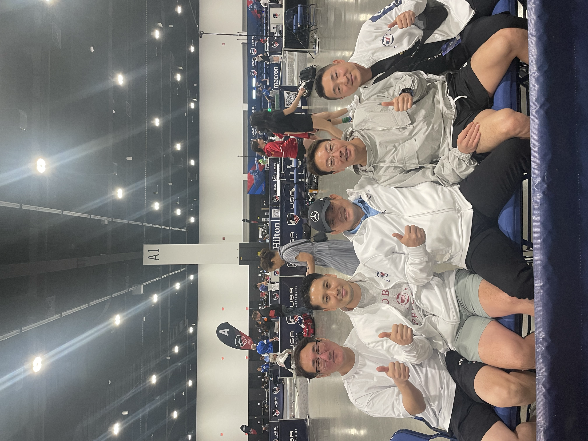 Four men sitting on chairs behind a table at an indoor competition, all smiling and giving thumbs up. The background shows a sports arena with people, banners, and screens, indicating an event related to US Soccer.