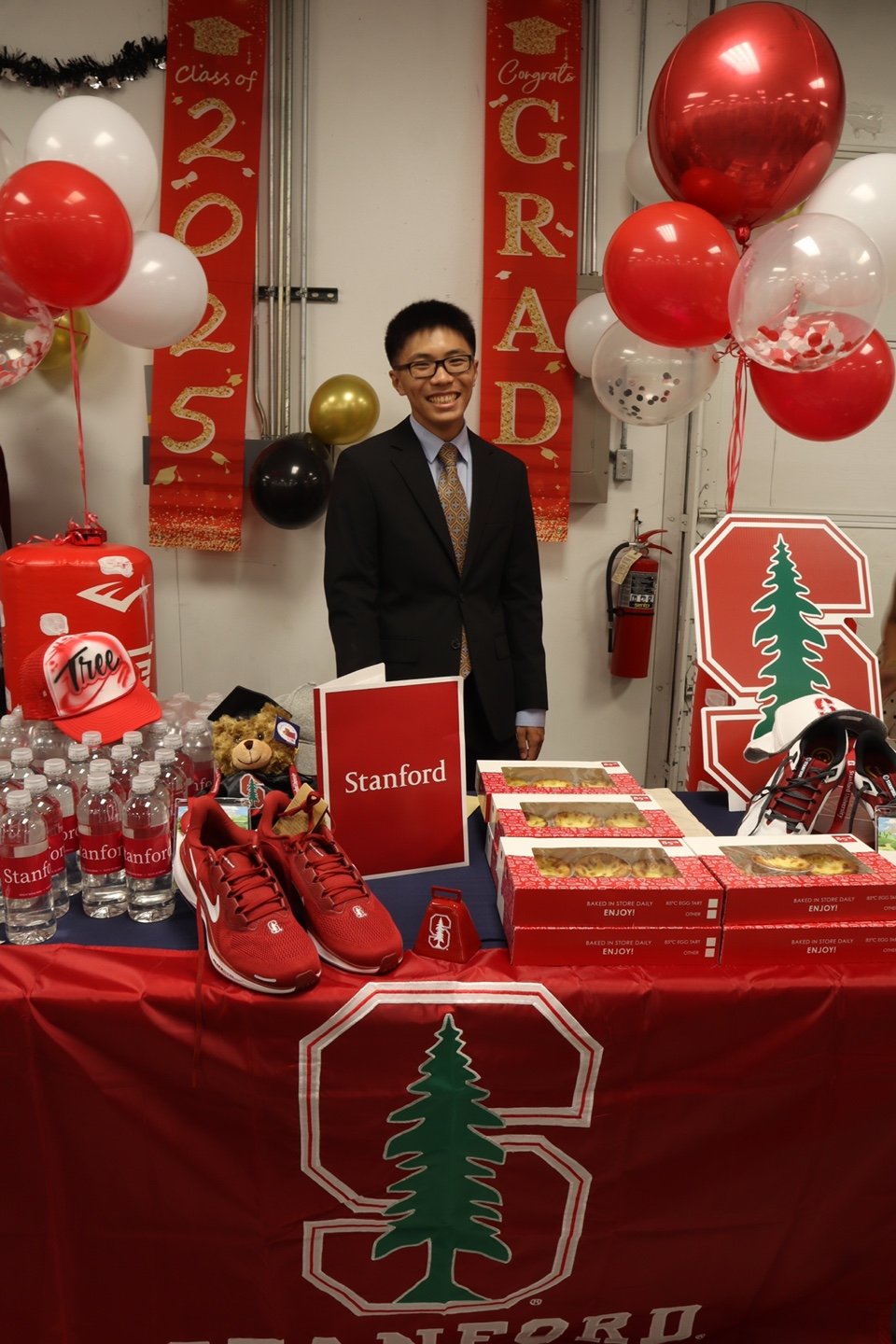 A young man in a suit and tie standing behind a table decorated with Stanford University items. The table has water bottles, pizza boxes, a teddy bear, and a pair of red Nike shoes. Banners behind him display 'Class of 2025' and 'Congrats GRAD.' Ball