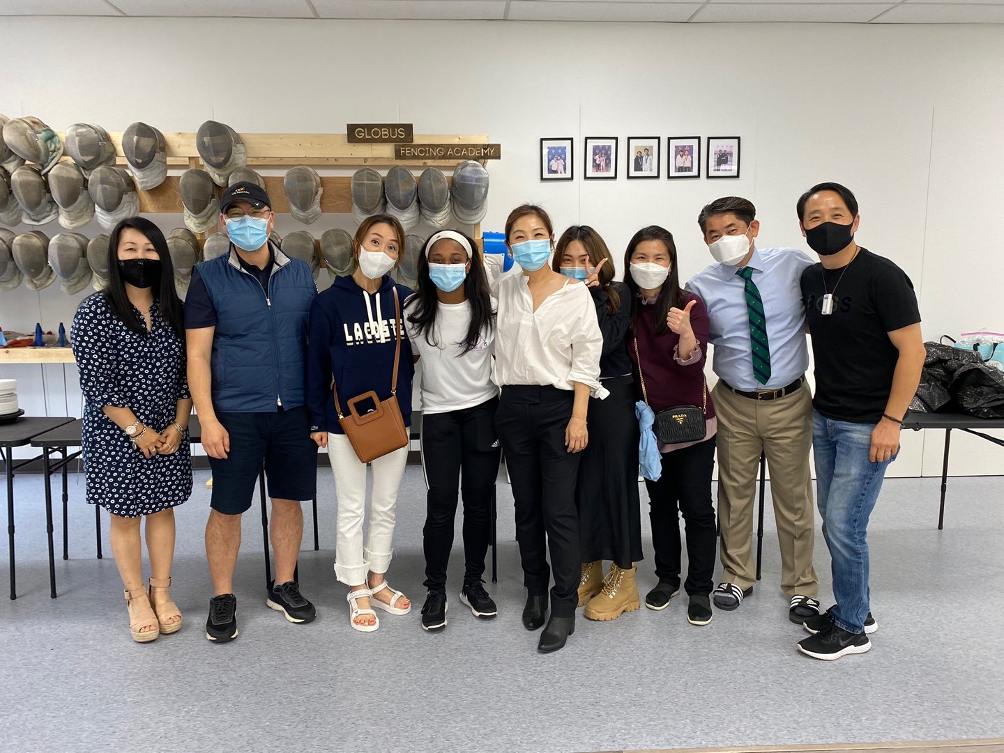 Group of nine people standing indoors, wearing masks, posing for a photo, with fencing helmets on the wall behind them and framed photos on the white wall.