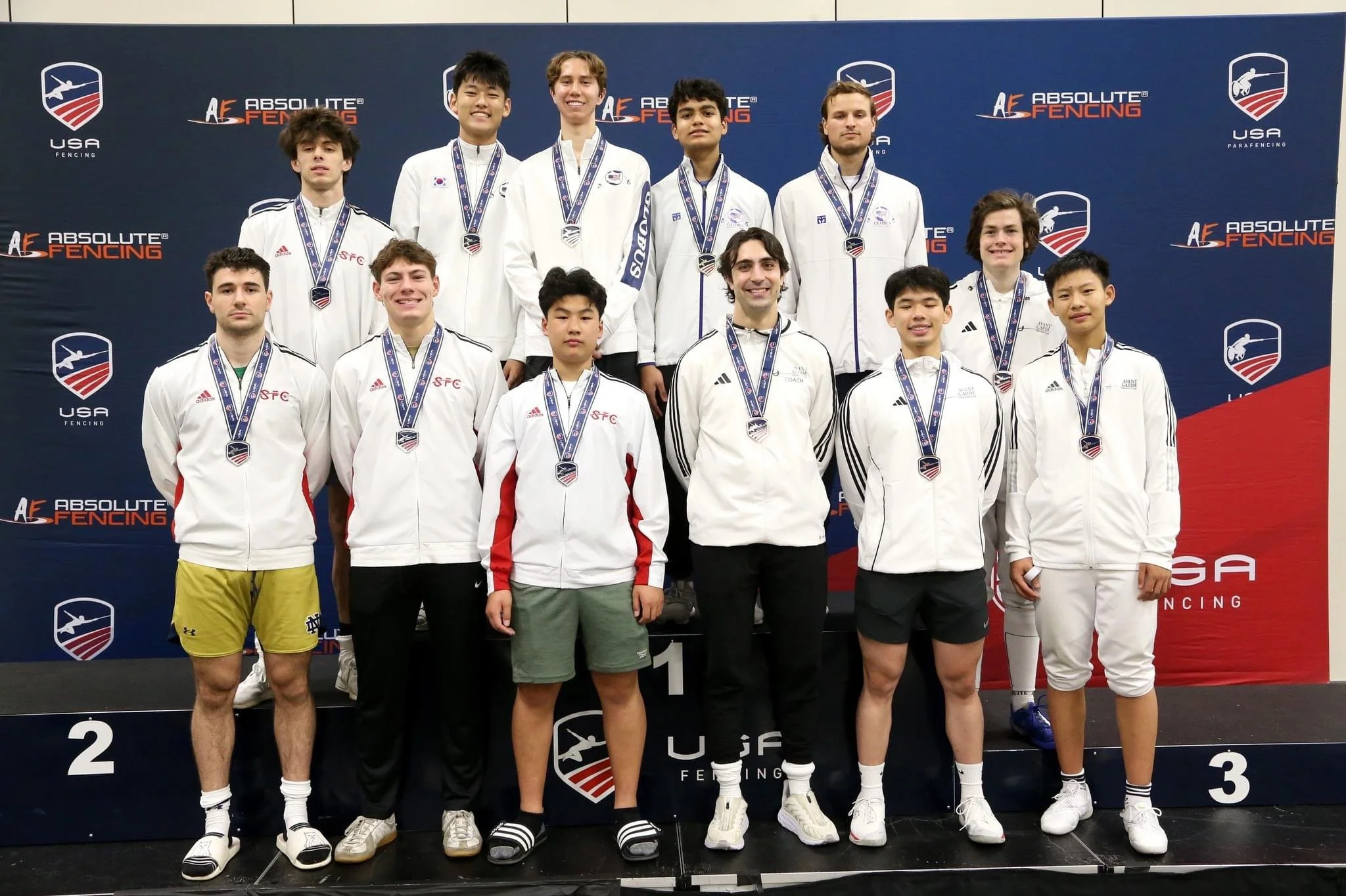 A group of young athletes standing on a podium with medals, celebrating after a fencing competition. They are dressed in sports jackets and shorts, with a backdrop displaying the logos of USA Fencing and Absolute Fencing.