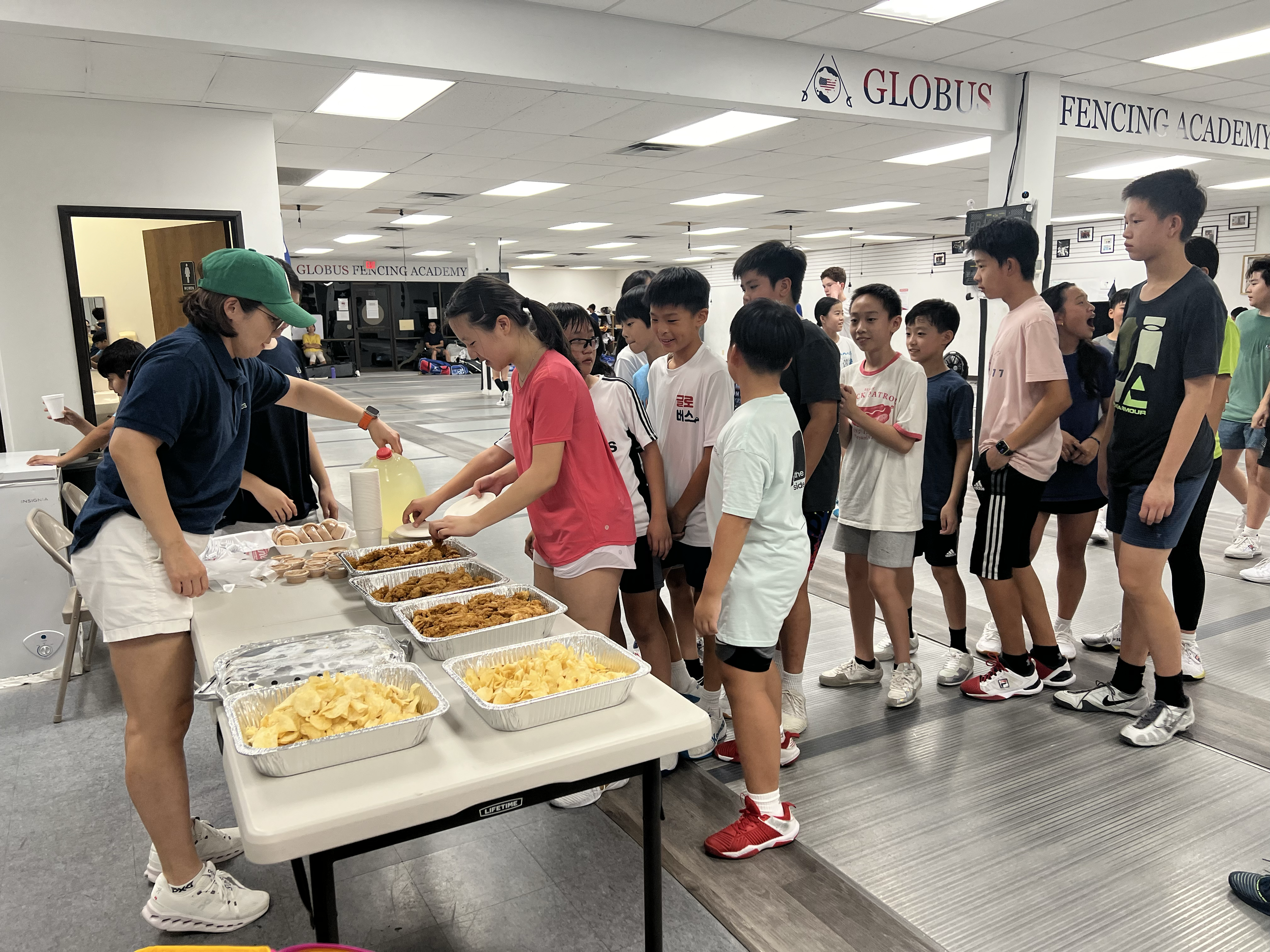 A group of children in line at a food table with fried chicken, potato chips, and drinks inside a fencing academy.