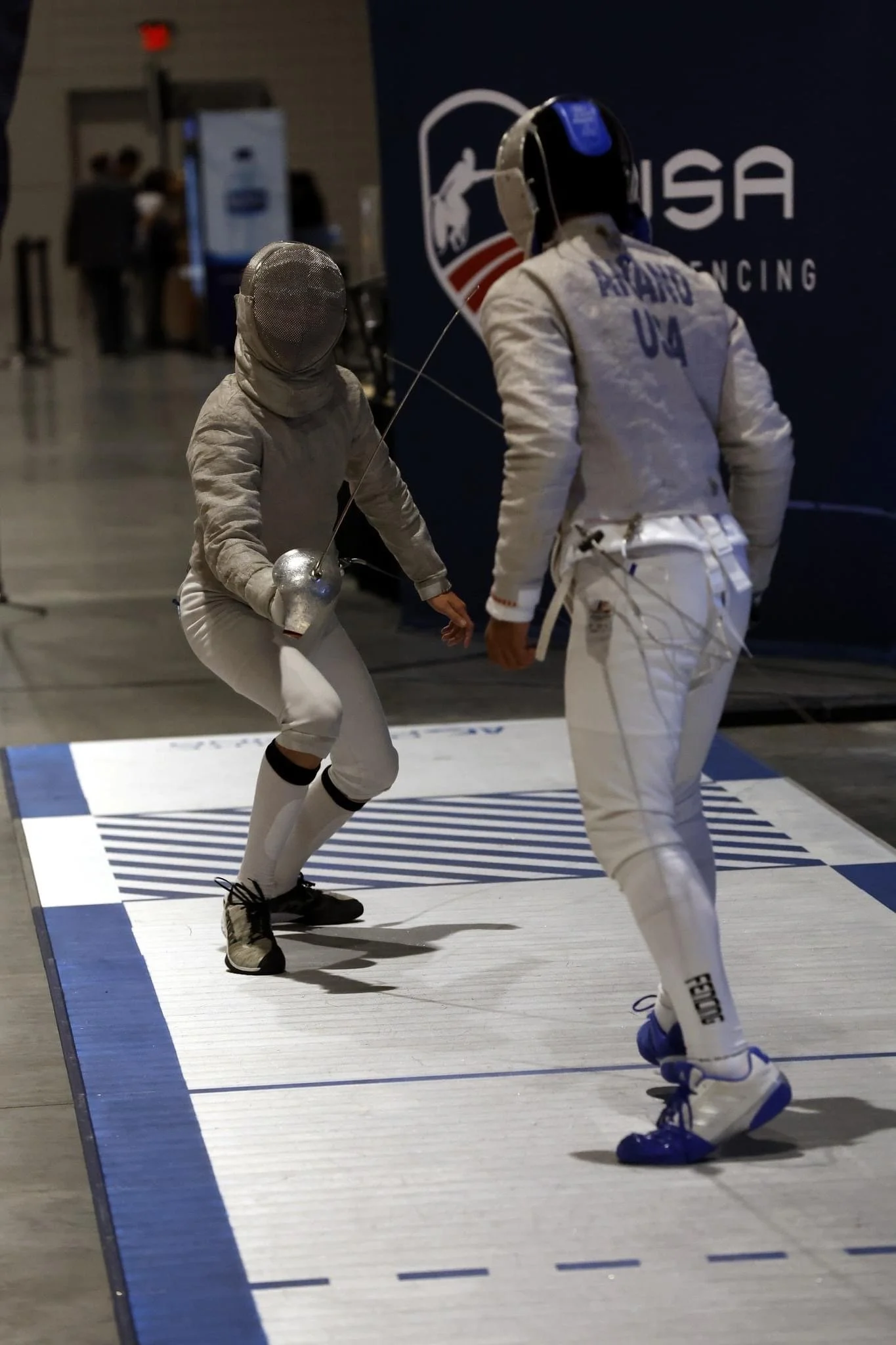 Two fencers face off on a fencing piste, one near the camera and the other in the background, both dressed in protective gear and masks, with the United States Fencing logo visible on the background wall.