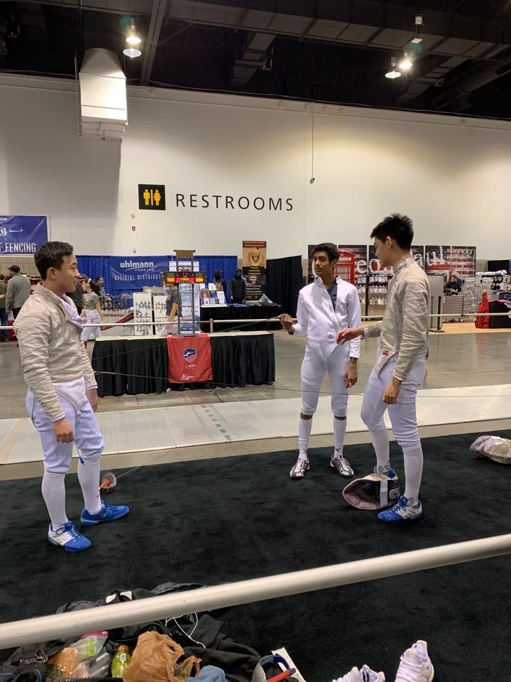 Three young men in fencing gear standing in a circle, talking, in an indoor sports facility with a sign that reads 'Restrooms' in the background.