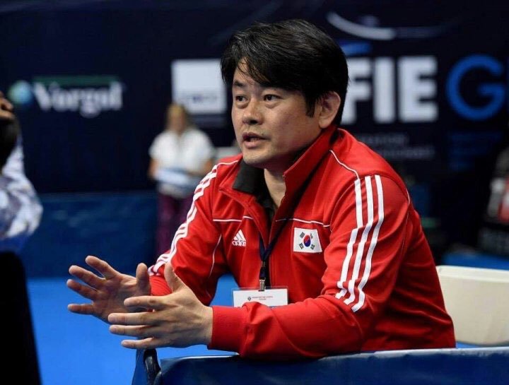 A man wearing a red South Korea sports jacket with white stripes and a South Korea flag on the chest, sitting at a table during what appears to be a sports event or competition.