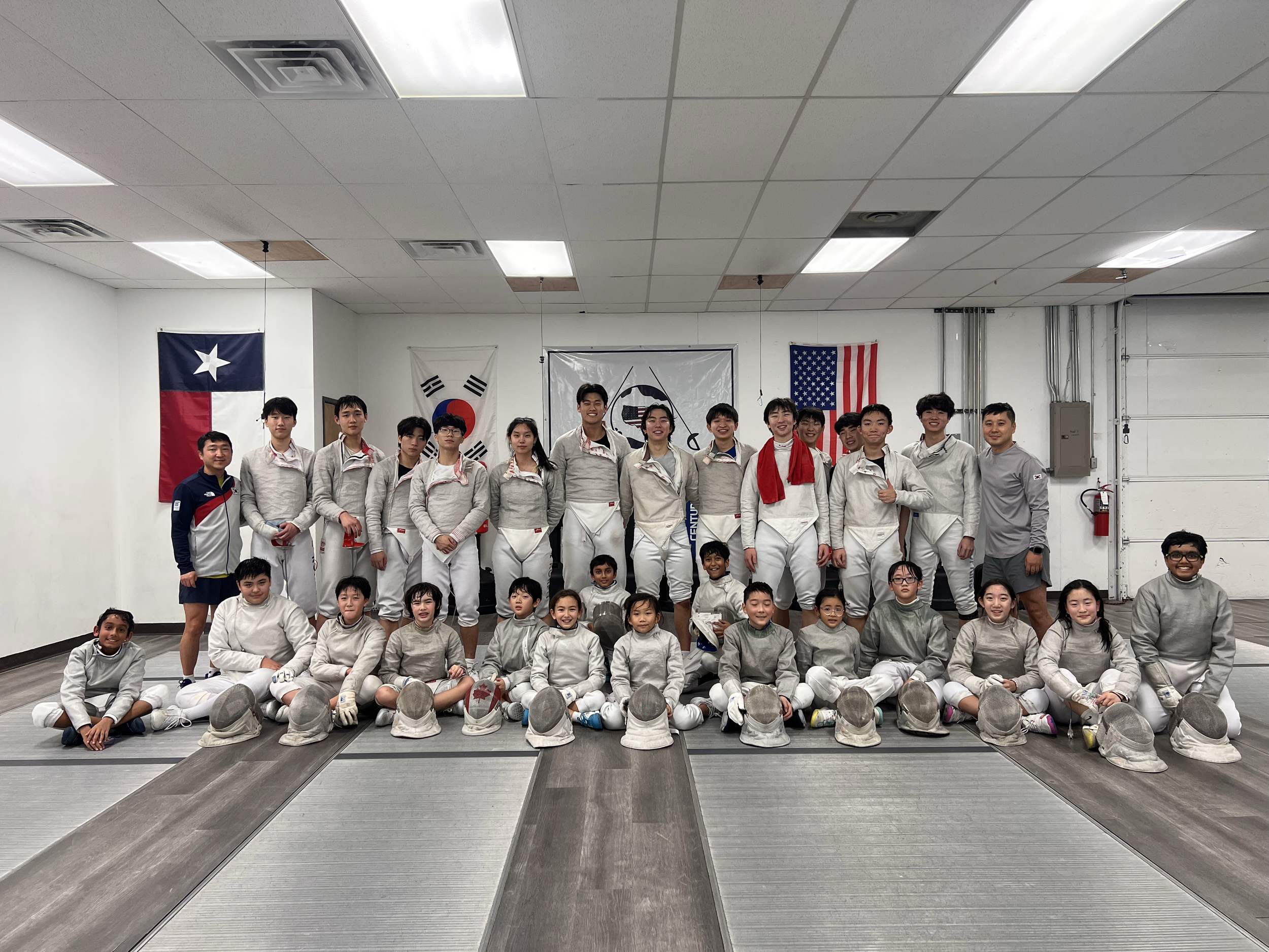 Group of young people in fencing gear, posing in a sports training facility. Flags of Texas, South Korea, and the United States are hanging on the wall behind them.