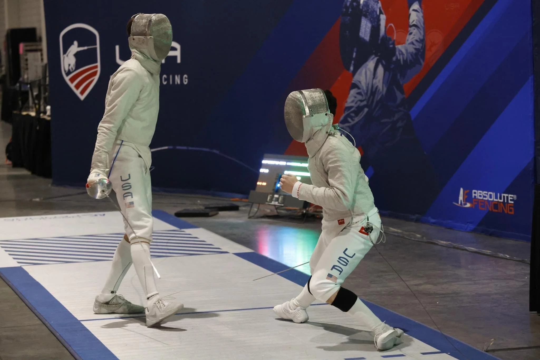Two fencers in white gear and masks fencing on a padded strip at an indoor sports event.