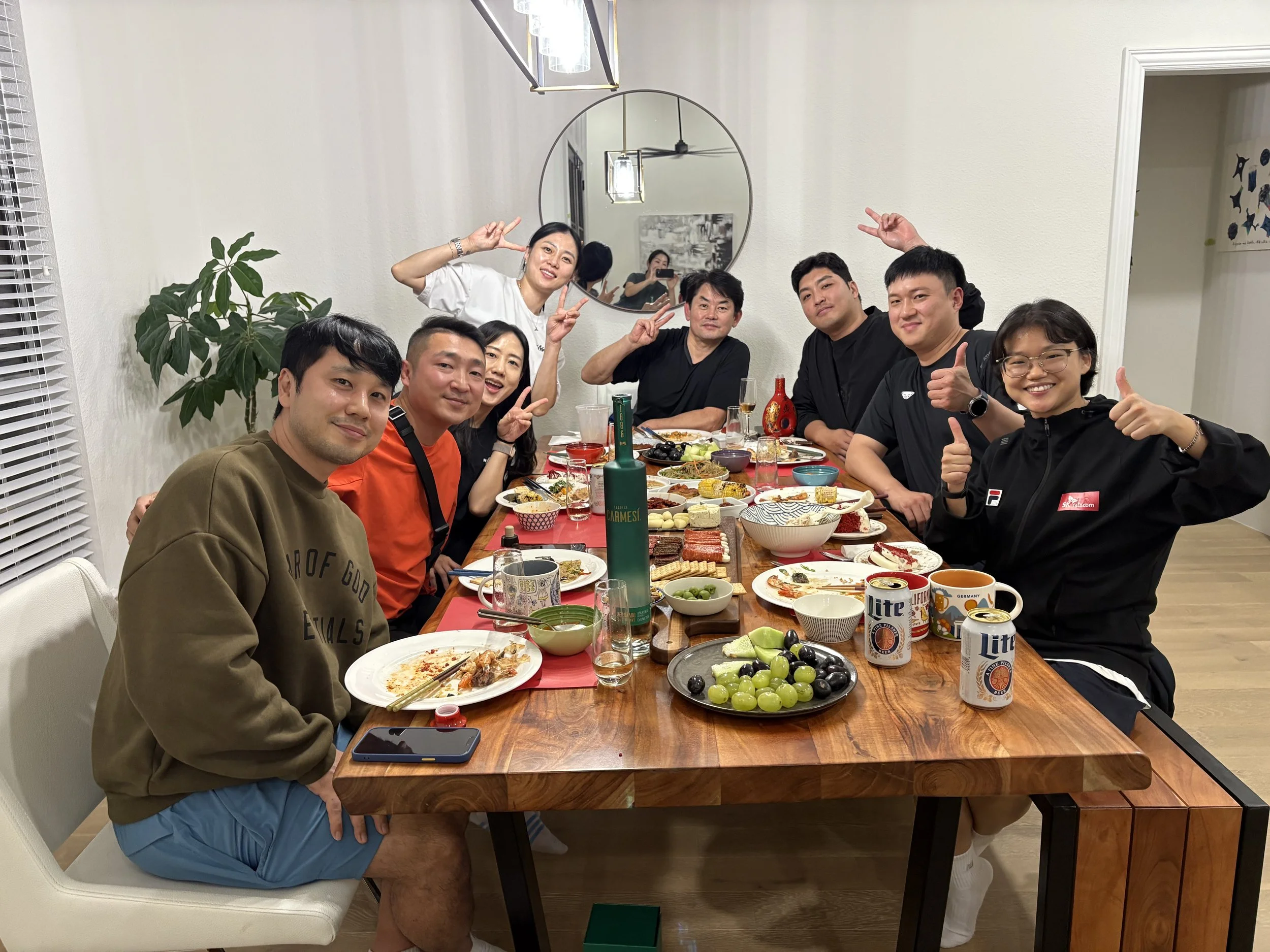 A group of nine people sitting around a dining table filled with food and drinks, smiling and making peace signs or thumbs-up gestures, in a well-lit dining room with a large mirror on the wall and a plant in the corner.