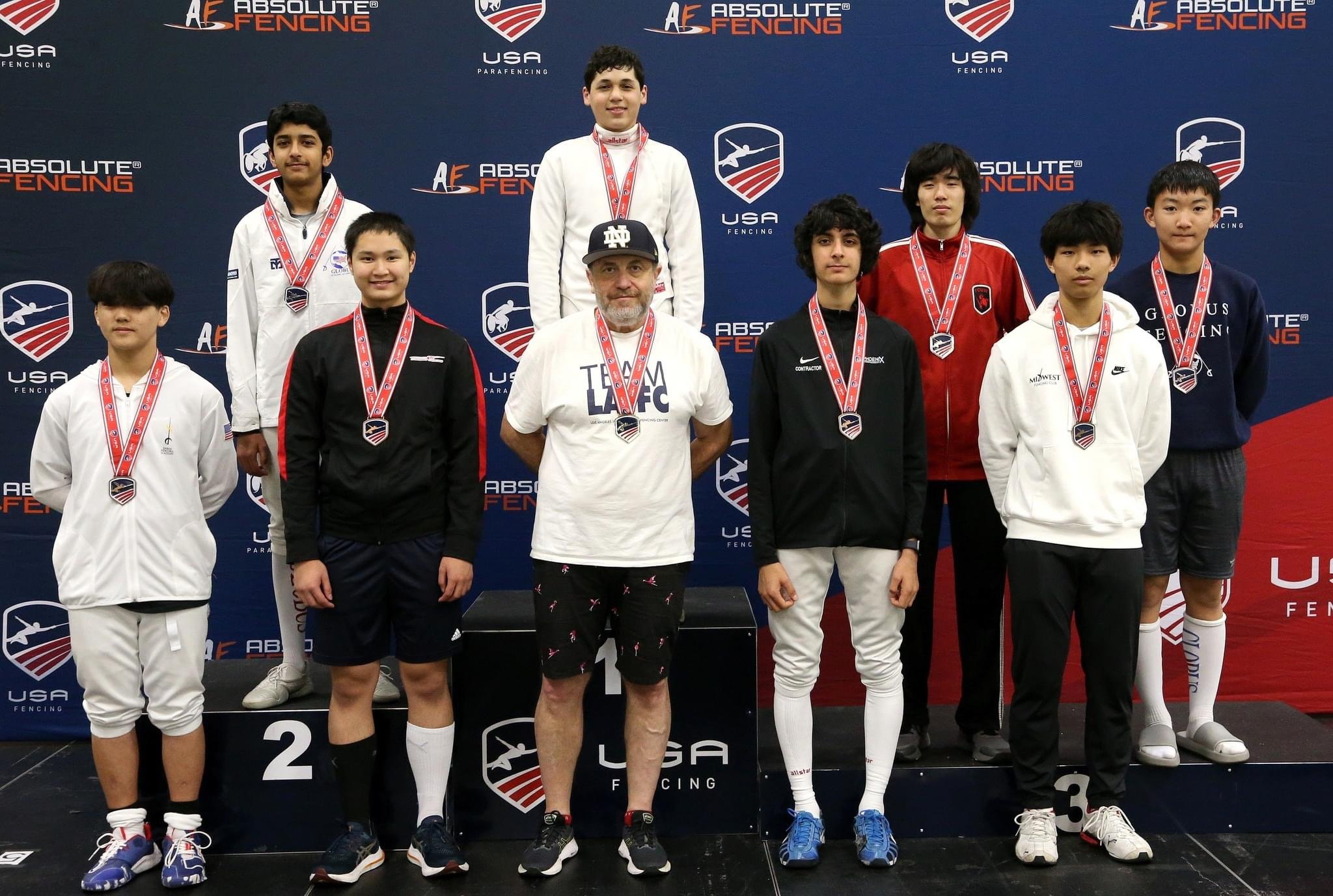 A group of young male athletes and one coach standing on a winners' podium at a fencing competition, with medals around their necks, in front of a blue backdrop with USA Fencing and Absolute Fencing logos.