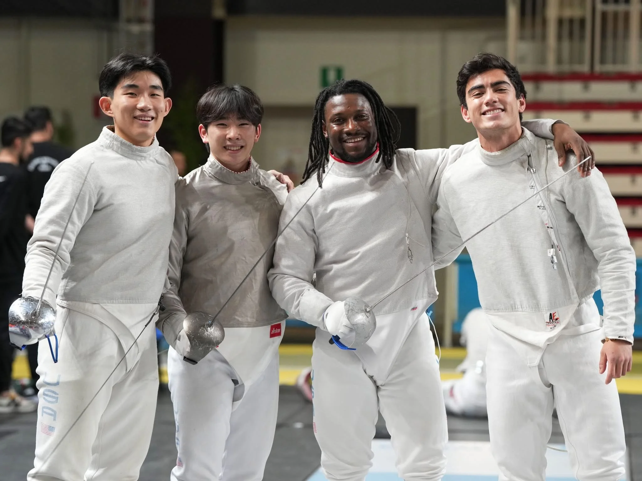 Four male fencers standing together, smiling, in fencing gear in a sports hall.