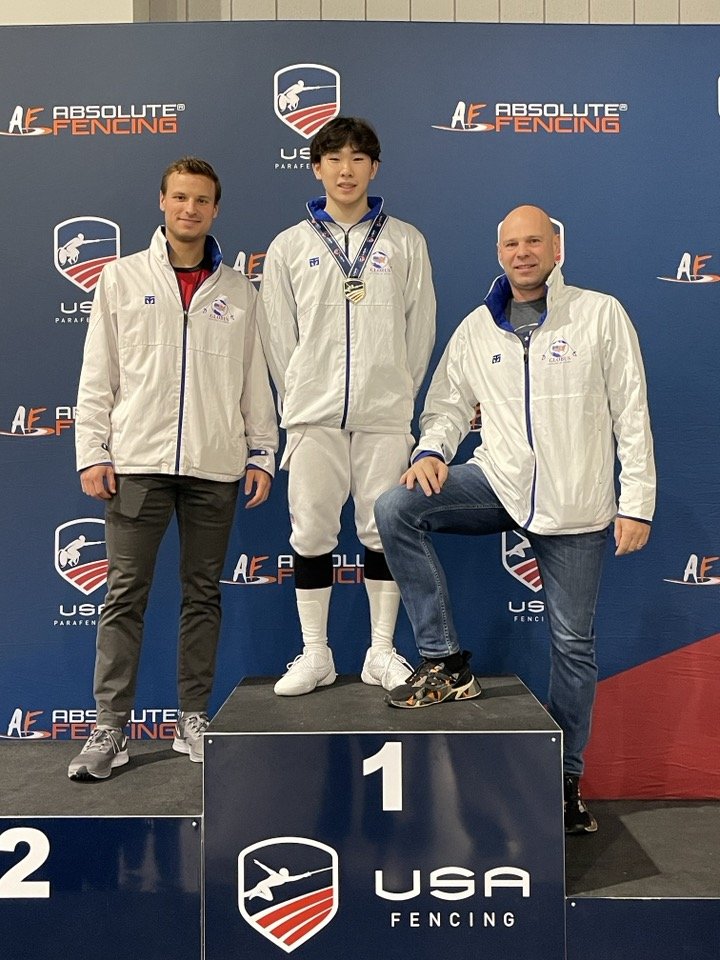 Young man standing on first place podium at a USA fencing competition, flanked by two men in white jackets, with medals around his neck, in front of a blue backdrop with USA fencing logos.