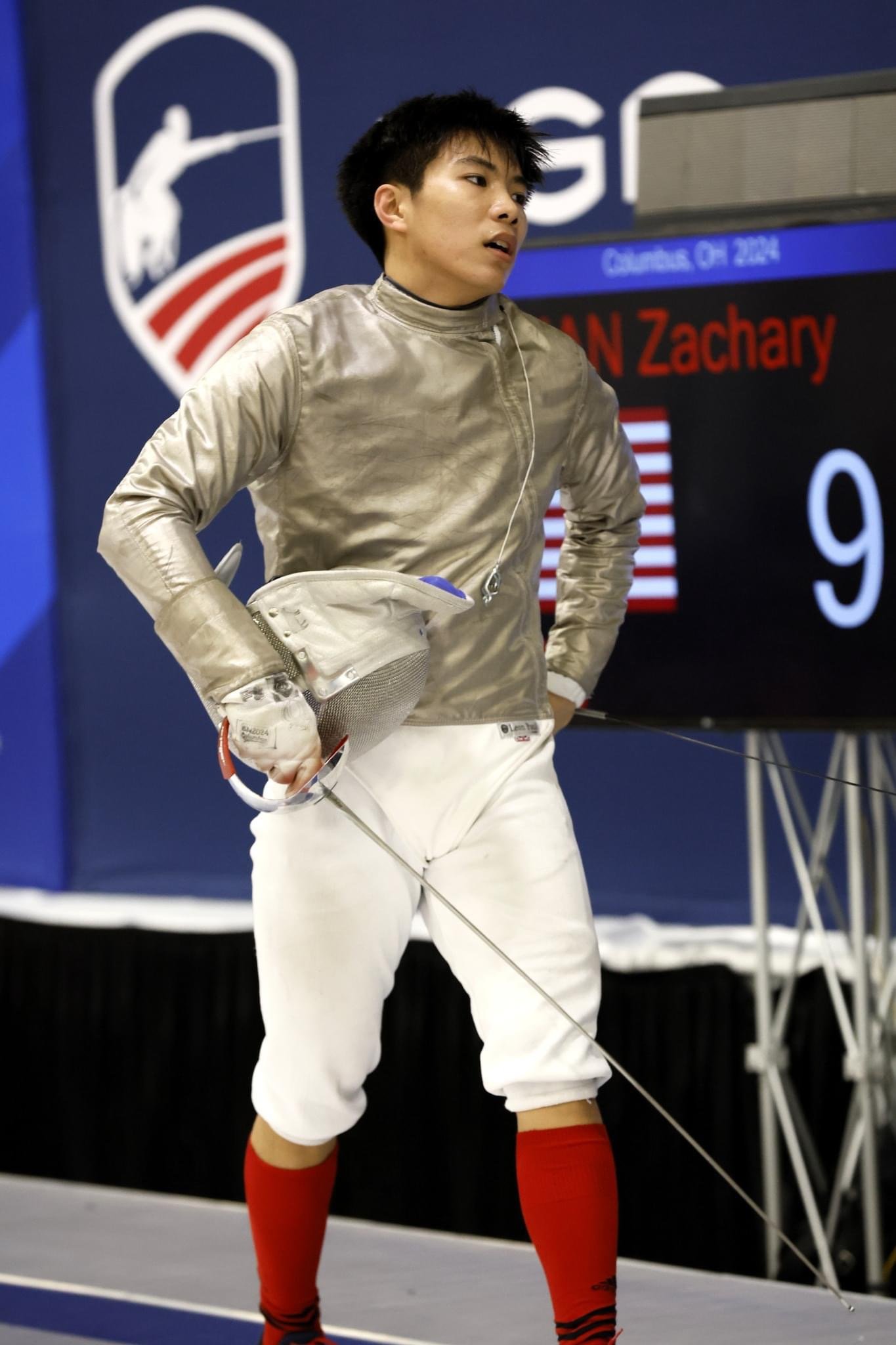 Young male fencer with short black hair in a silver fencing jacket, white fencing pants, and red knee-high socks standing with his hand on his hip, holding a fencing mask and weapon, in an indoor sports arena with scoreboard and fencing logo in the b
