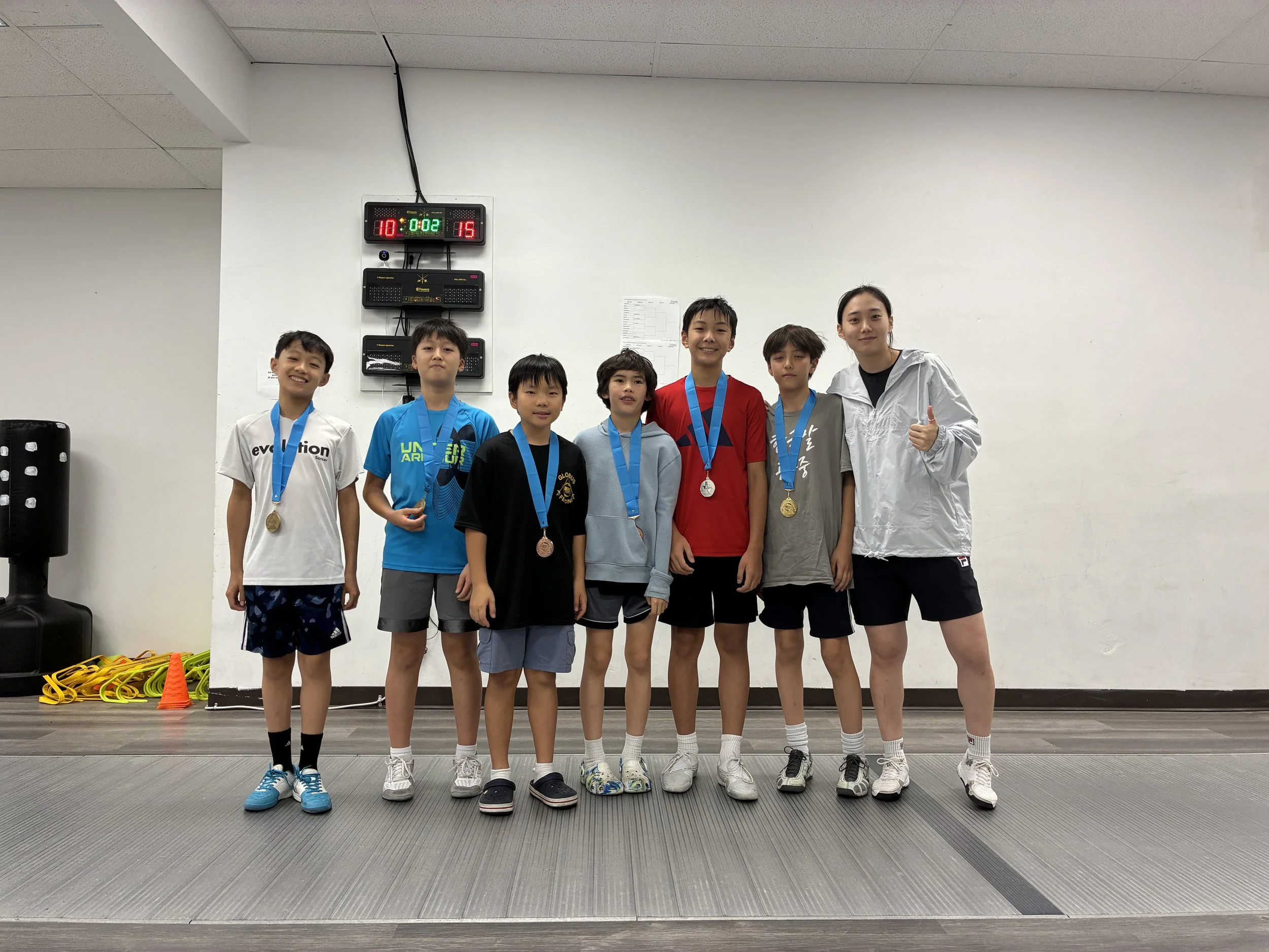 Group of seven young boys and girls in sportswear standing on a mat with medals around their necks, inside a gym or sports facility, with a scoreboard and equipment on the wall behind them.