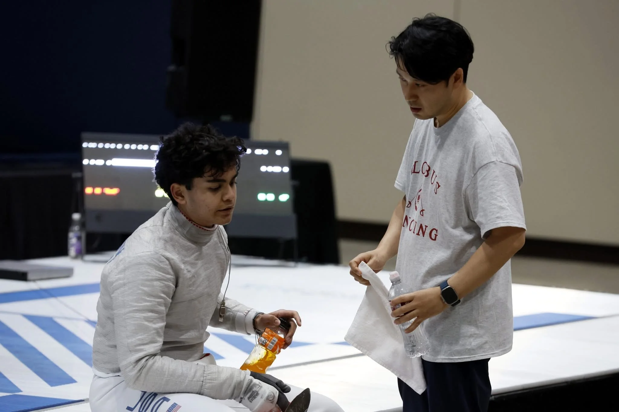 A young male fencer receiving water and snacks from a man in a casual t-shirt during a break at a fencing competition.