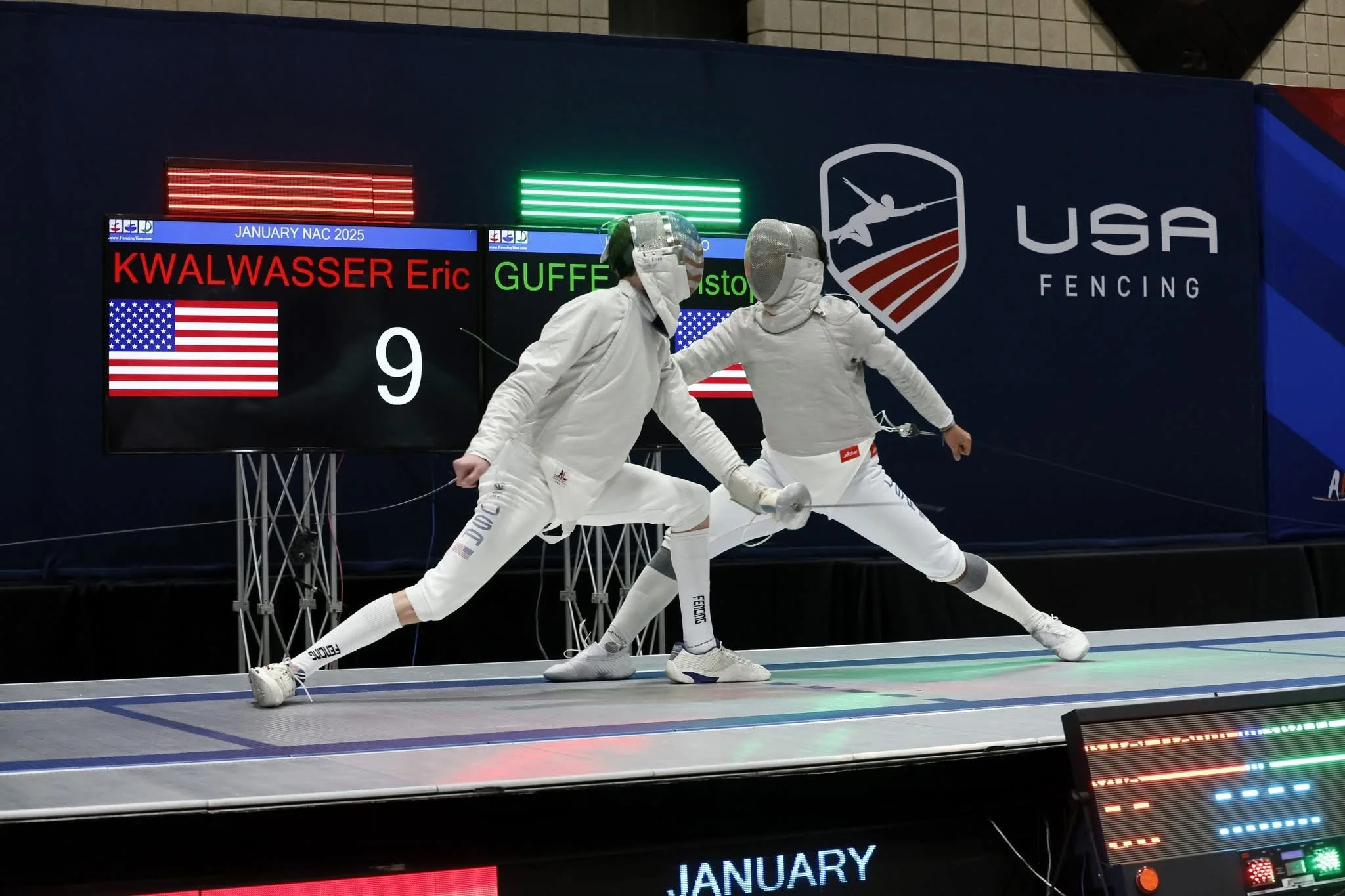 Two fencers compete in a match at a USA Fencing event, dressed in white protective gear, on an electronic fencing strip, with a scoreboard behind them showing their names and scores.