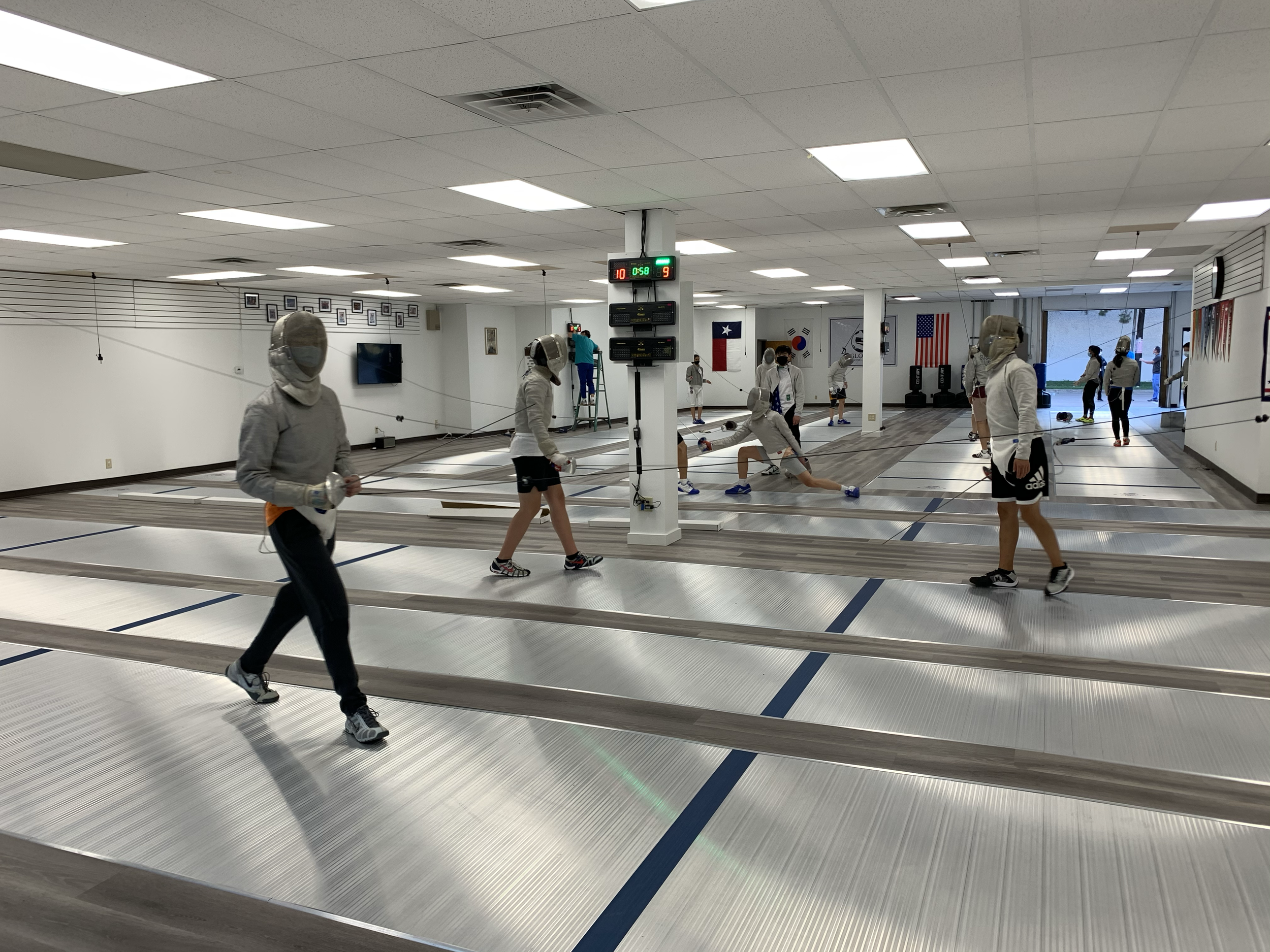 People practicing fencing in an indoor training facility, wearing protective gear and masks.