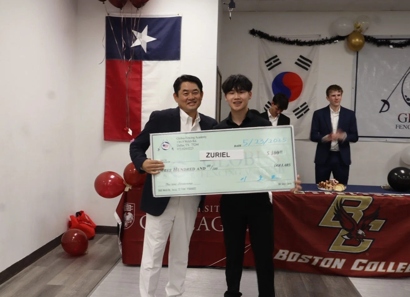 Two men standing in front of a table with a large check, one man is holding the check and the other is pointing to it. There are flags of Texas and South Korea hanging on the wall behind them, and balloons and decorations on the table.