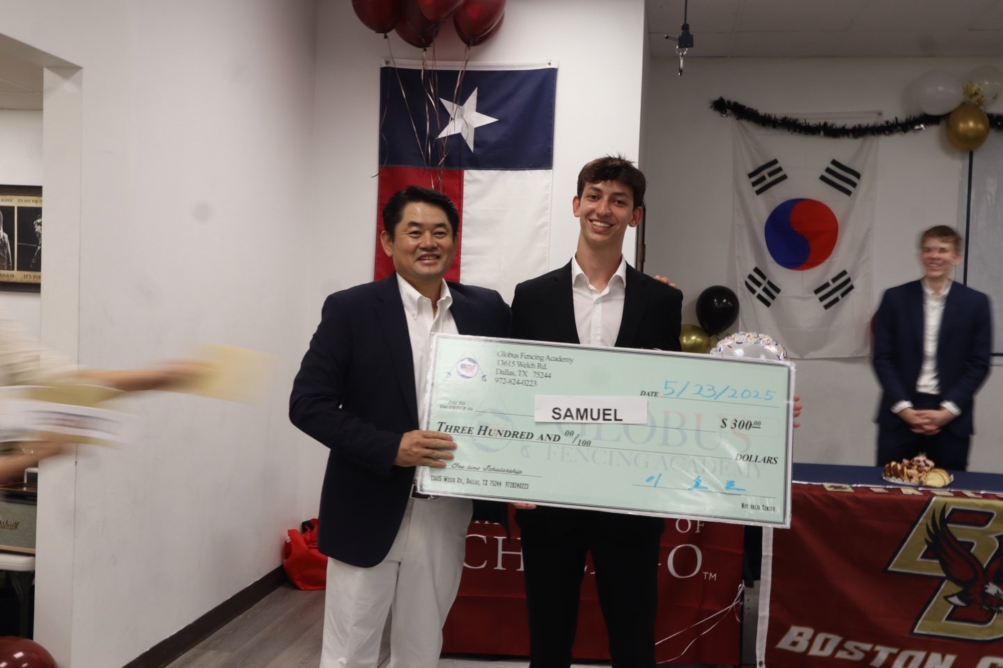 Two men in suits holding a large check with a young man. The background shows flags, including the Chilean and South Korean flags, and balloons at an indoor event.