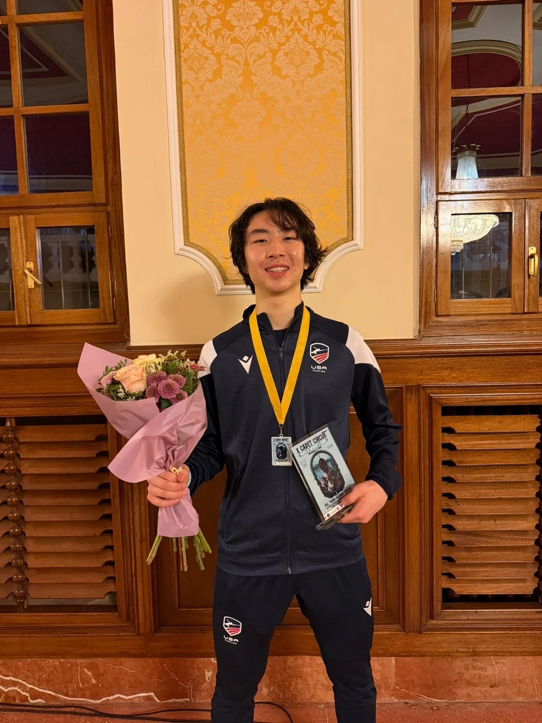 Young man in USA Olympic team jacket holding a bouquet of flowers and a medal and diploma, standing in front of a wooden-paneled wall with decorative wallpaper.