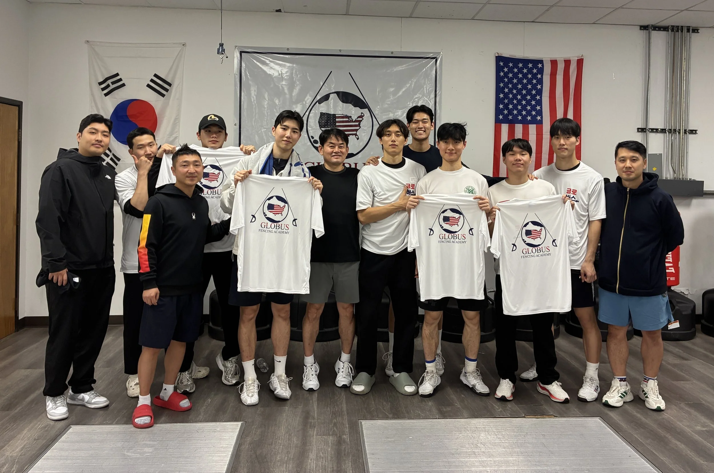 Group of 12 young men posing indoors, some holding 'GLOBUS FENCING ACADEMY' T-shirts, with South Korean and American flags in the background.