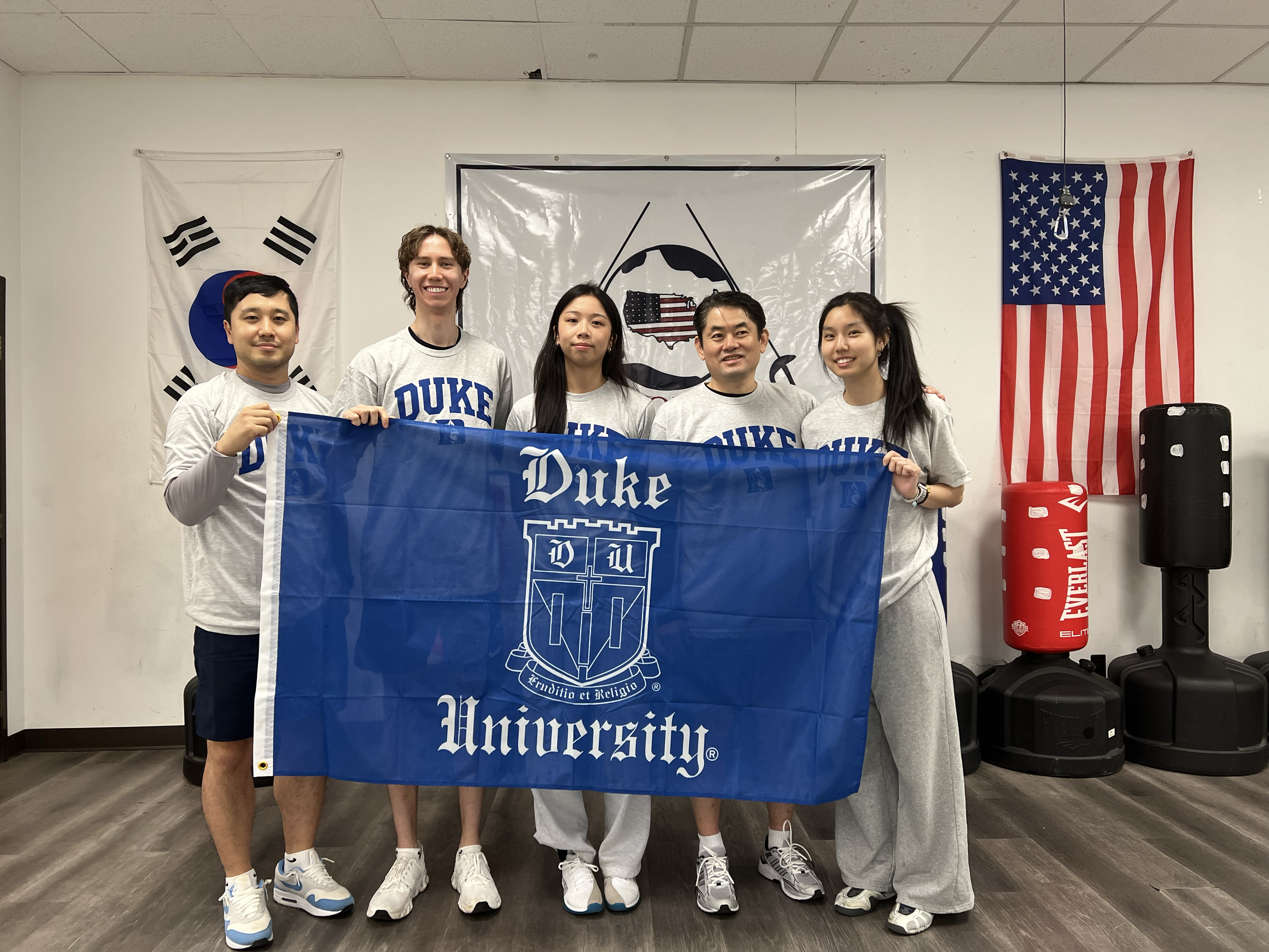 Group of five people holding a blue Duke University banner, standing in front of American and South Korean flags, in an indoor setting.