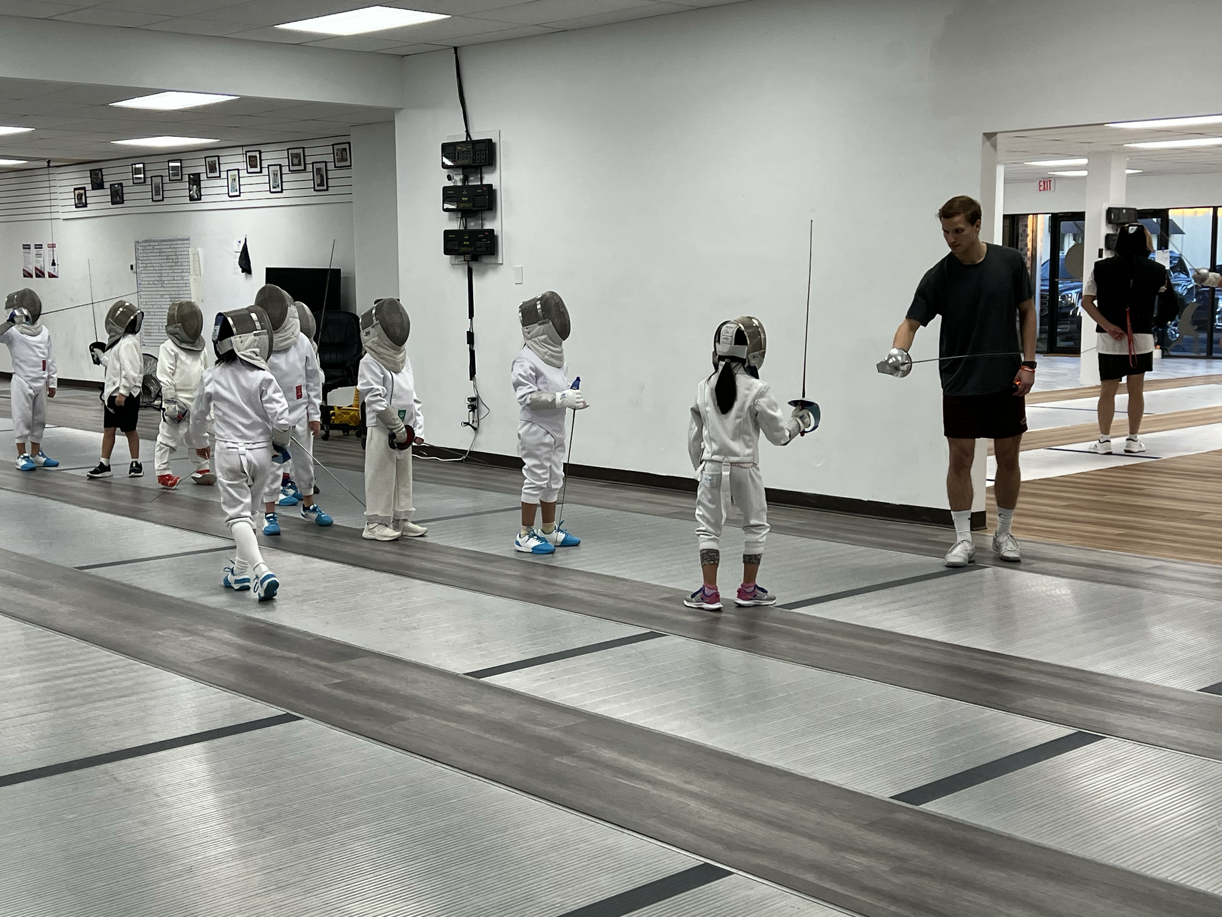 Children in fencing protective gear practicing fencing with two coaches in an indoor sports facility.