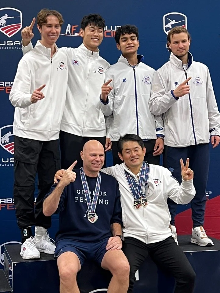Group of six male athletes, wearing white and blue sports jackets with medals around their necks, celebrating on a podium with USA Para Fencing logos and banners in the background.