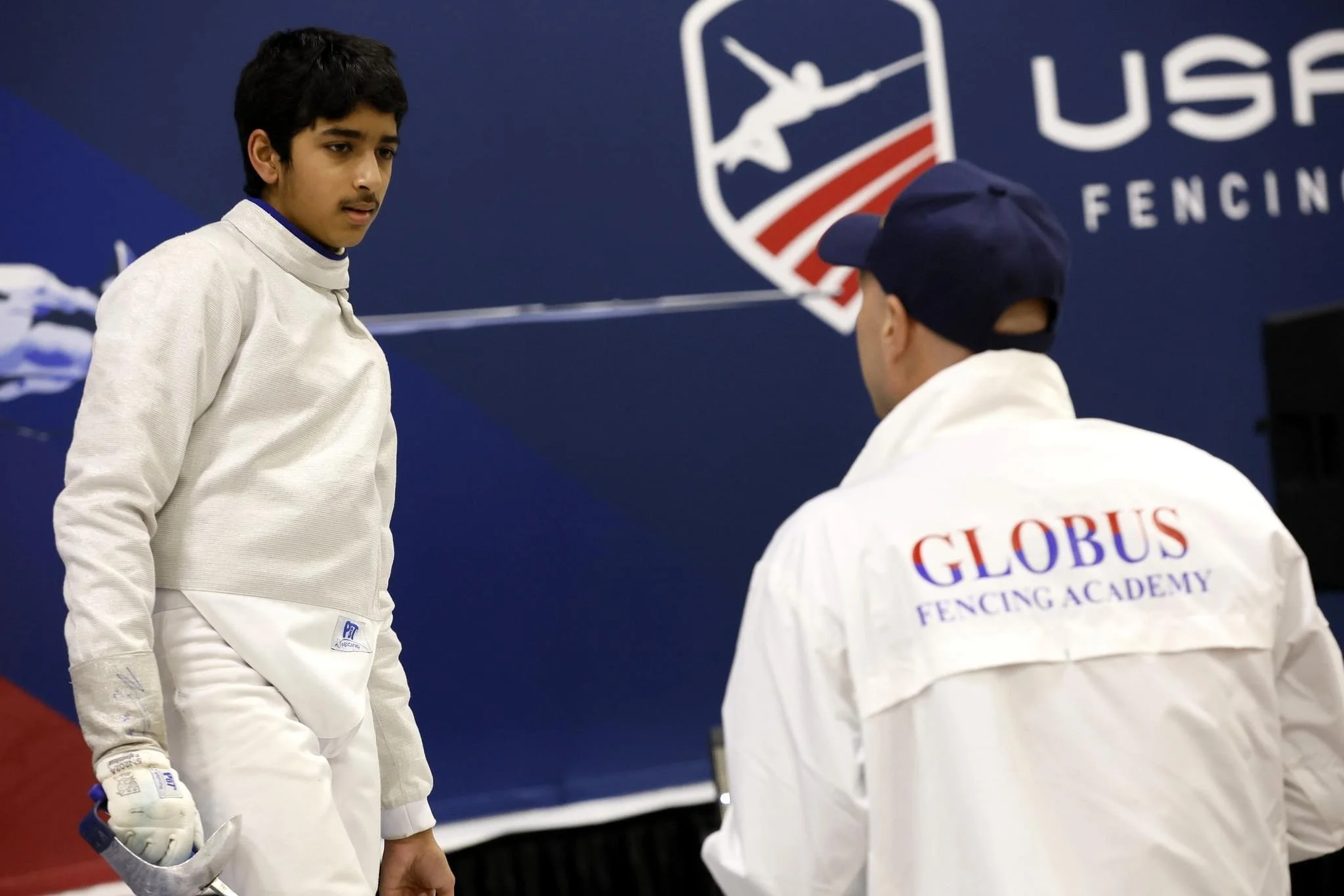 A young male fencer in white fencing gear stands during a match or practice, holding a fencing foil, while a coach or teammate in a jacket with "GLOBUS FENCING ACADEMY" on the back talks to him. There is a USA Fencing logo on a blue background behind