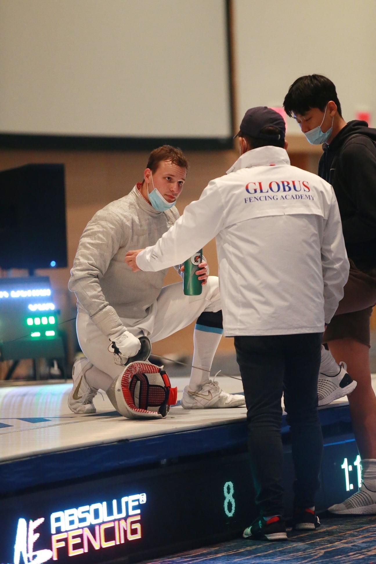Fencer wearing white uniform kneeling on fencing strip while talking to her coach, who is wearing a white jacket with 'GLOBUS FENCING ACADEMY' on the back. Two other individuals are standing nearby, all wearing face masks.