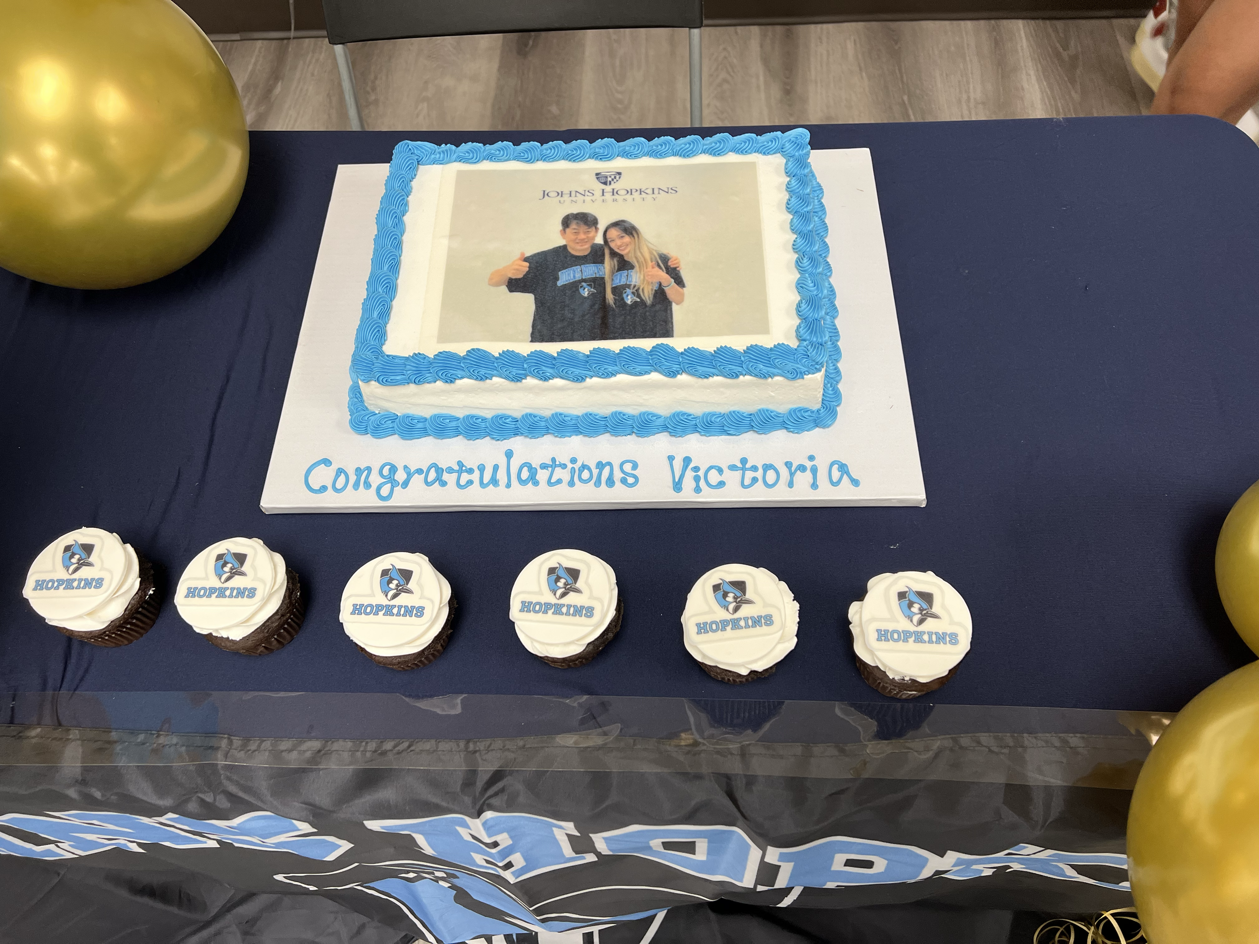 A graduation cake with a photo of two graduates, blue piping, and the text "Congratulations Victoria". There are also six cupcakes with the Hopkins logo, and gold balloons around the table.