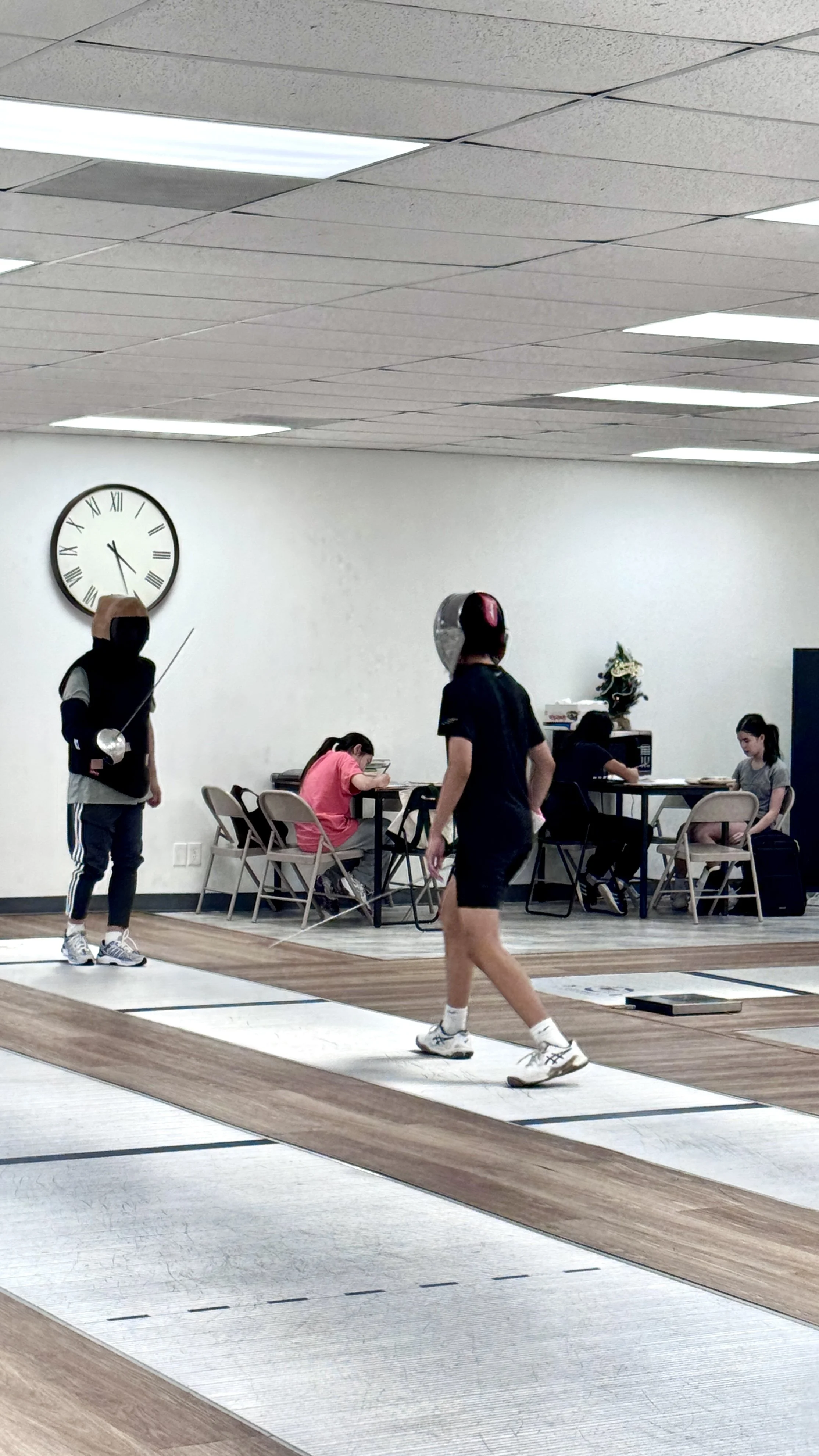 Two people practicing fencing indoors on a designated fencing strip with fencing gear, including masks and foils.