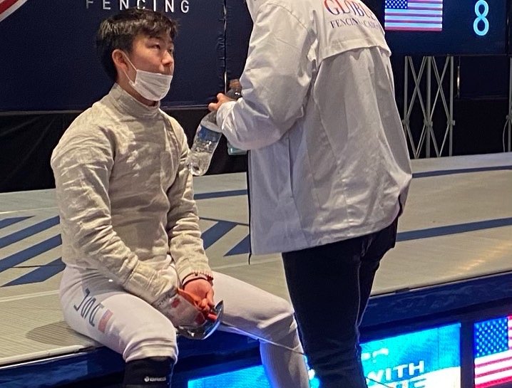 A young fencer sitting on a bench and talking to an adult, both wearing fencing gear and masks, in an indoor fencing arena.