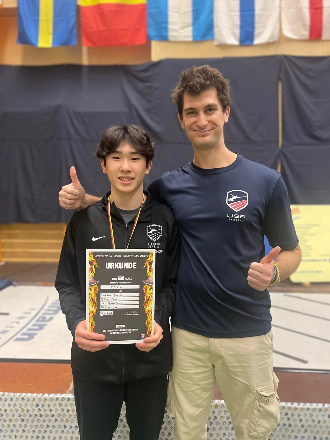 Two men standing side by side indoors, smiling at the camera, with flags hanging in the background. The young man on the left is holding a certificate and wearing a medal, while the man on the right is giving a thumbs up.