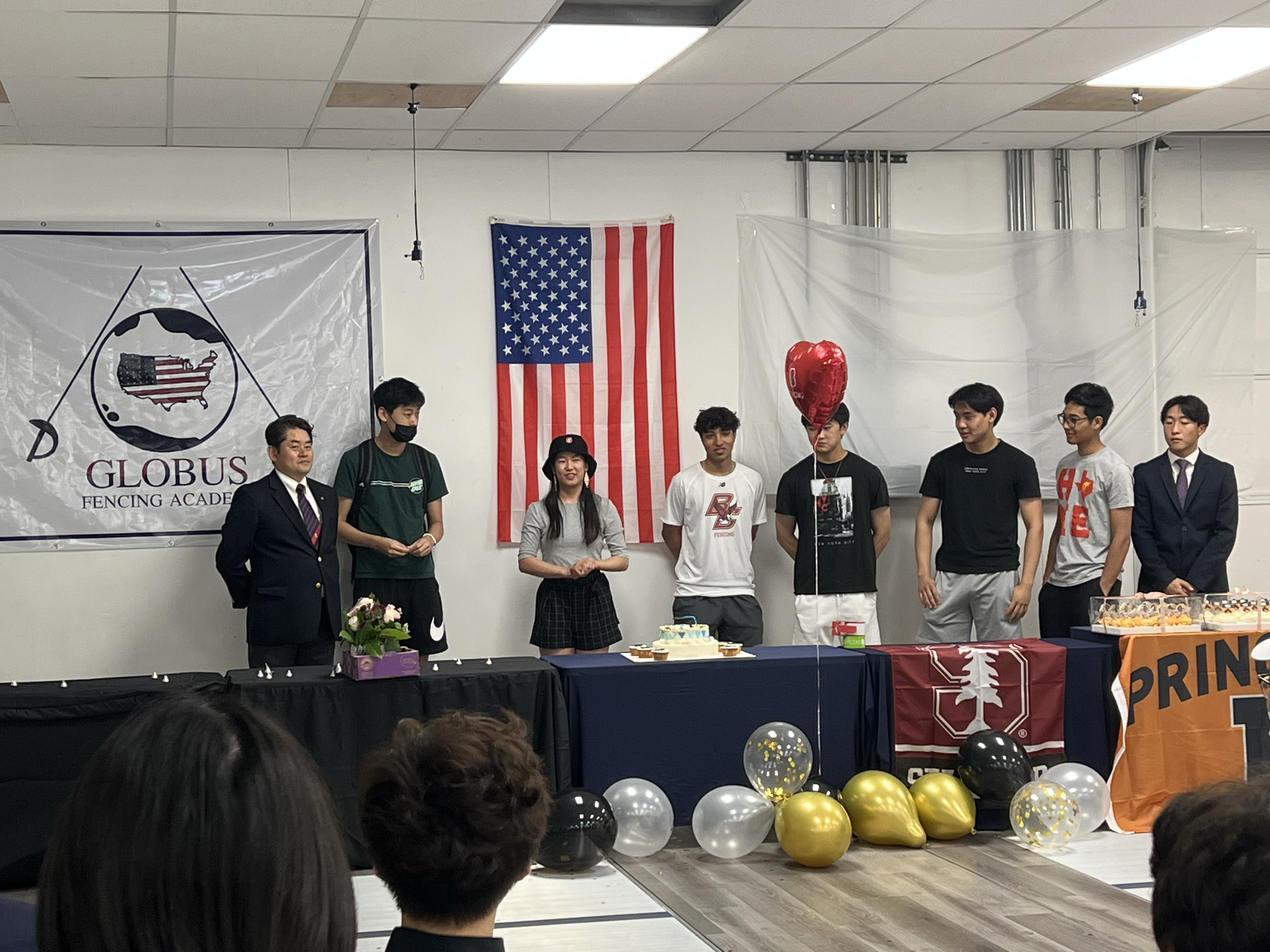 Group of people standing behind decorated tables with balloons, cake, and gifts at an indoor celebration, including a man in a suit, students, and American flags.