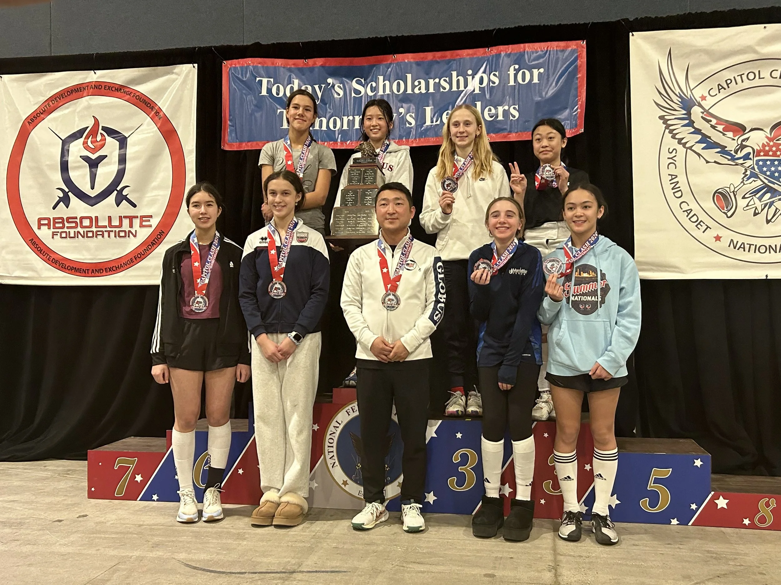 A group of young female athletes and one adult male standing on a winners' podium at a fencing competition, holding medals and trophies, with banners behind them displaying logos and text celebrating the event.