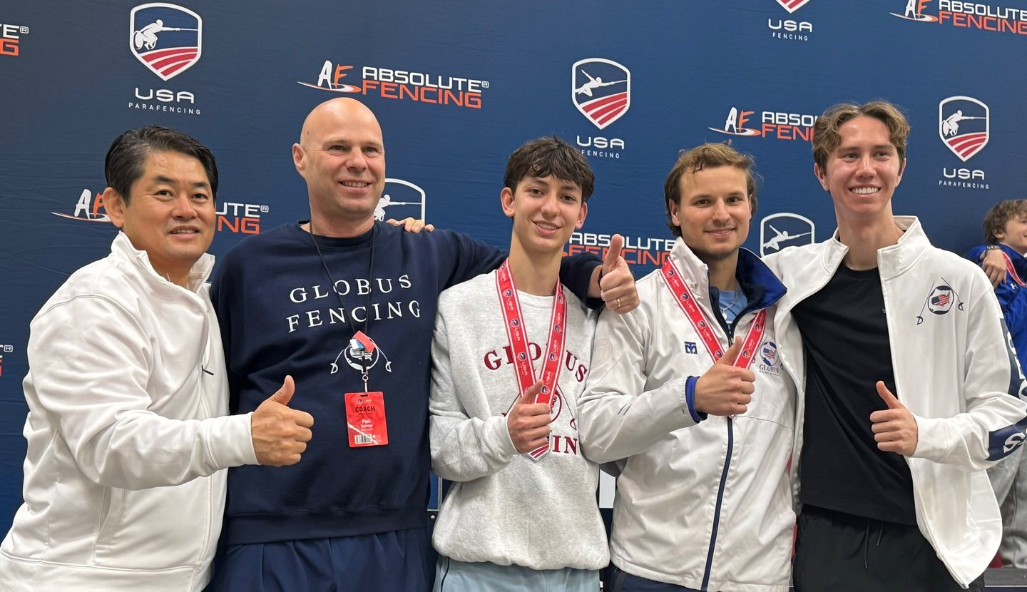 Group of five male fencers standing in front of a blue backdrop with logos and text. They are smiling, giving thumbs-up, and wearing medals and sports gear.