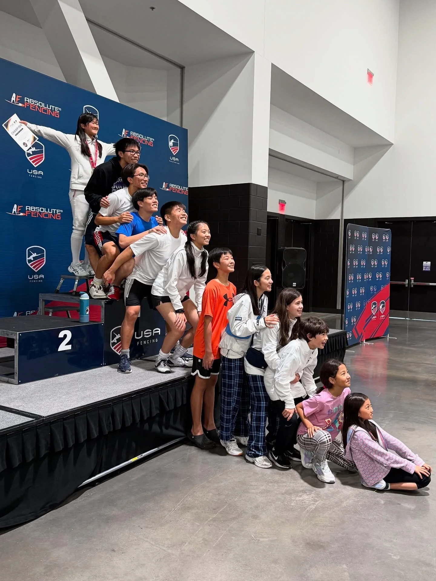Group of young athletes in fencing gear posing on a winners' podium at a USA Fencing event.
