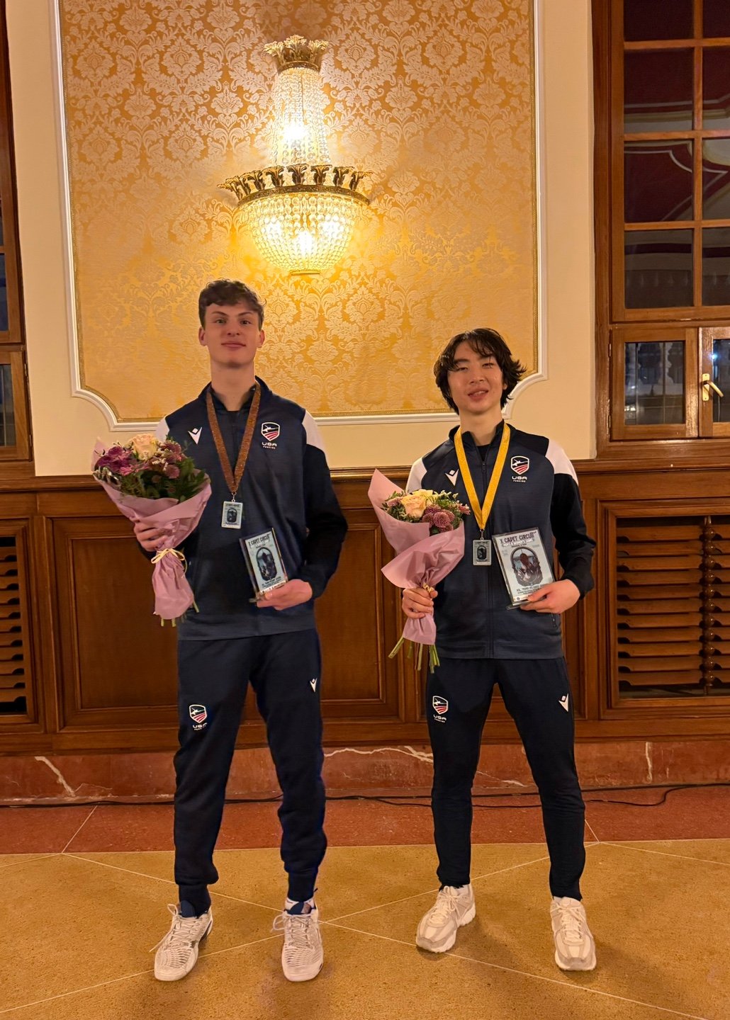 Two young athletes in matching USA team jackets holding bouquets of flowers and medals, standing in an ornate room with a chandelier and wooden paneling.