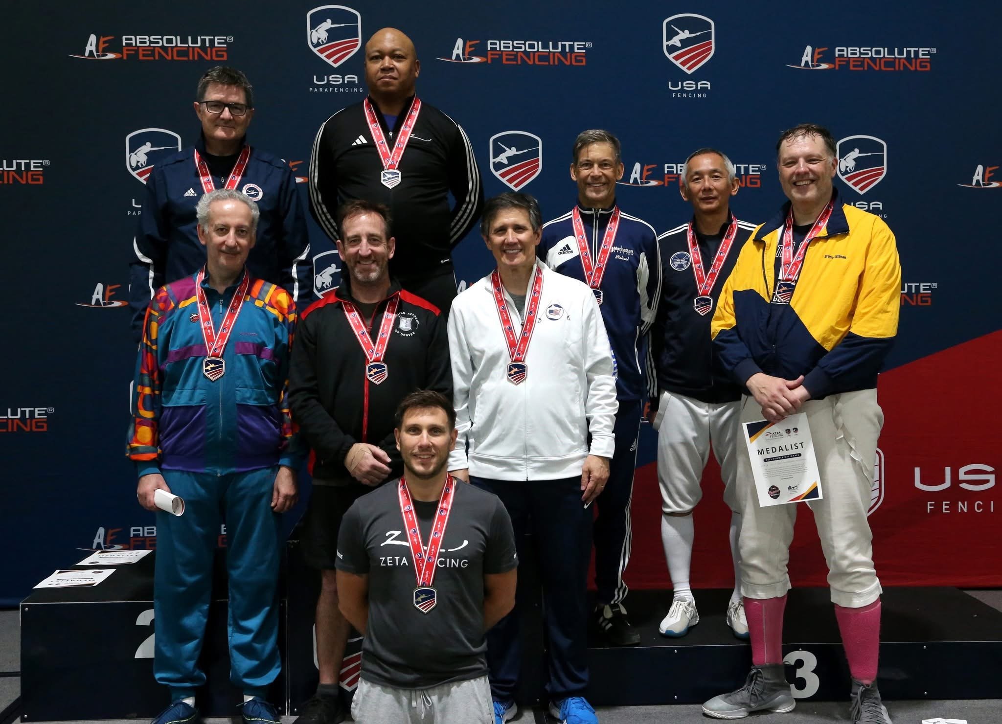 Group of nine male athletes celebrating on a winner's podium with medals and certificates, at an ABSOLUTE FENCING event in the USA, with a dark blue backdrop displaying logos and text.