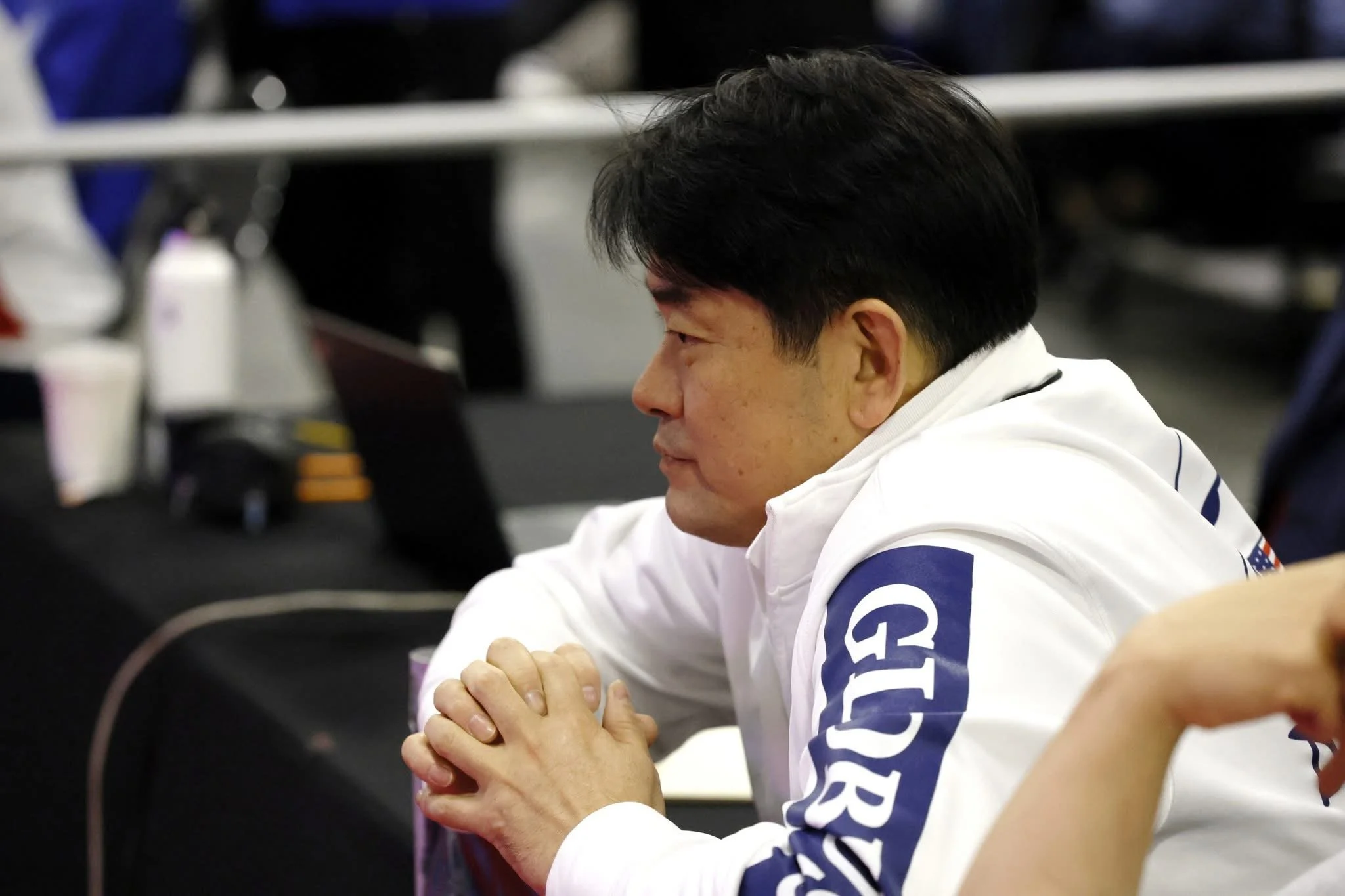 A man with black hair, wearing a white jacket with blue lettering on the sleeve, is sitting at a table with his hands clasped together, looking to his right.