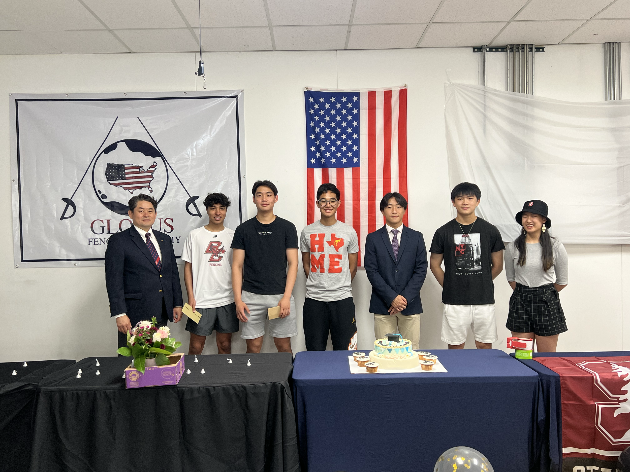 A group of seven people, including a man in a suit, standing behind a table with a birthday cake and cupcakes, in front of a U.S. flag and a banner with a logo for a fencing academy, in a decorated room.