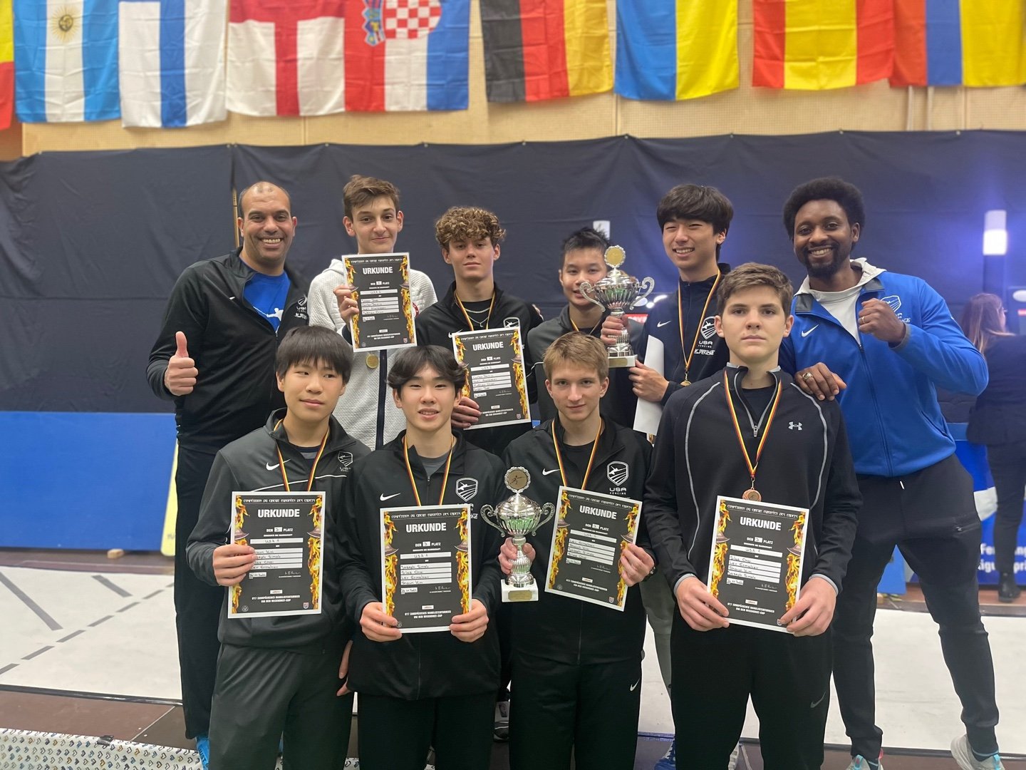 A group of young male athletes and their coaches posing indoors with medals, trophies, and certificates after winning a sports competition. Flags from various countries hang in the background.