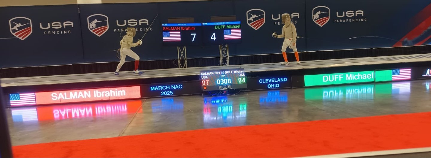 Two fencers practicing or competing in a fencing match at an indoor tournament with USA fencing branding. Digital scoreboards show the names Salman Ibrahim and DUFF Michael with scores 7 and 4. Fencers are wearing protective gear and holding their sw