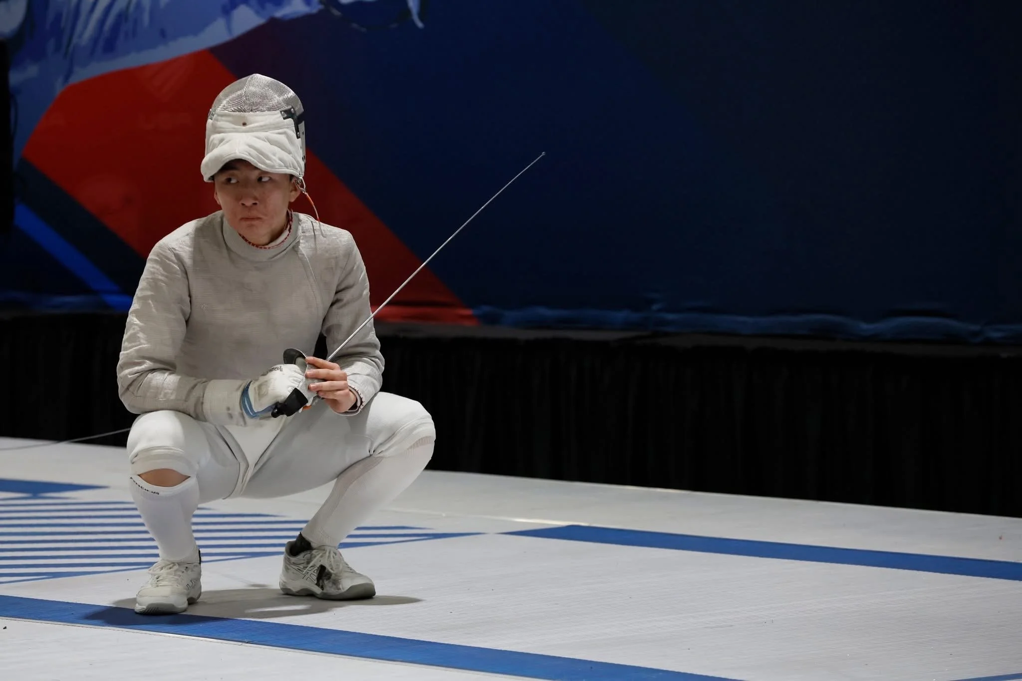 A female fencer squatting with a fencing sword in her right hand on a fencing piste.