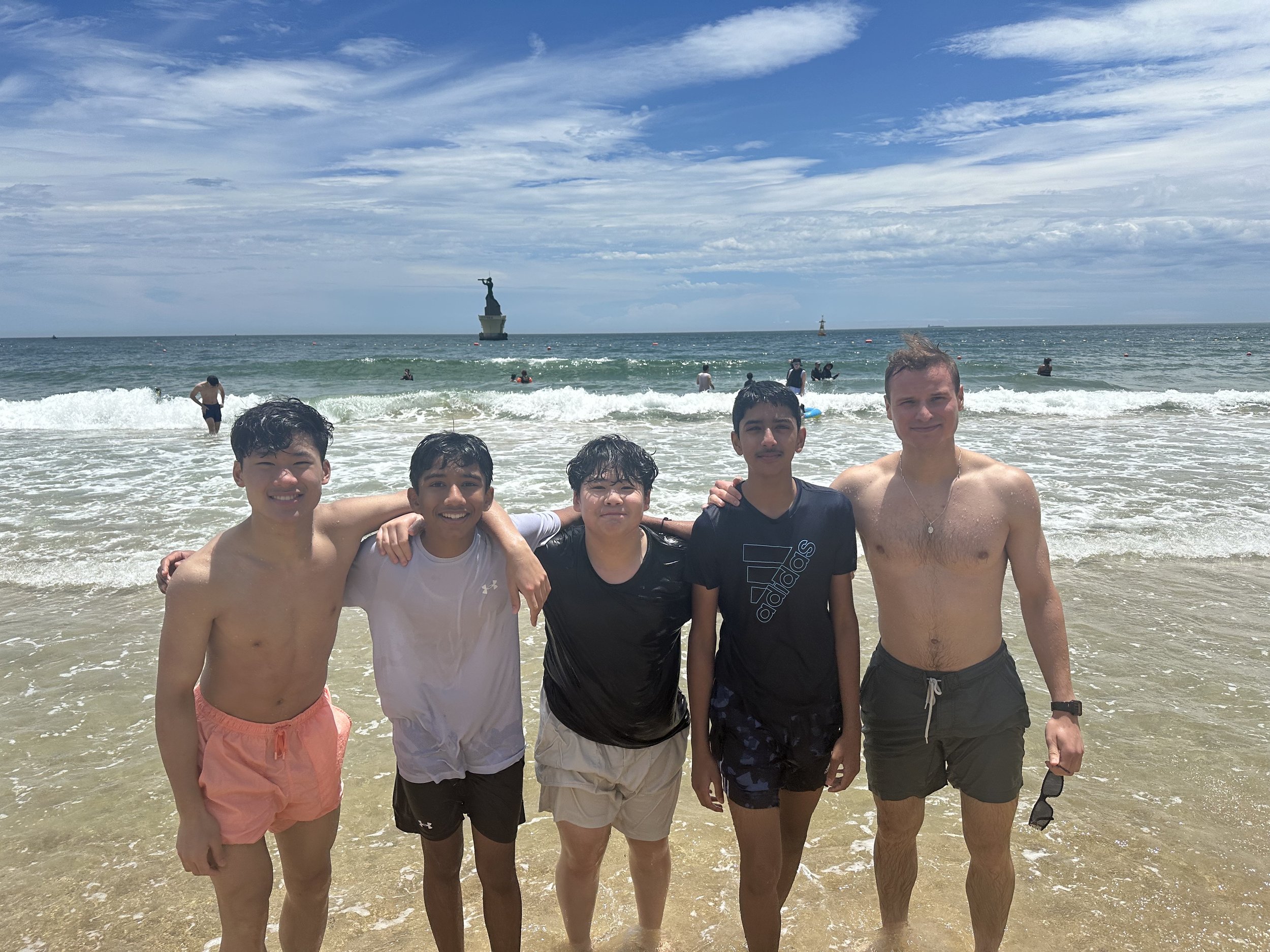 Five boys standing in the ocean with umbrellas, water, a lighthouse, and a statue on a boat in the background during a sunny day