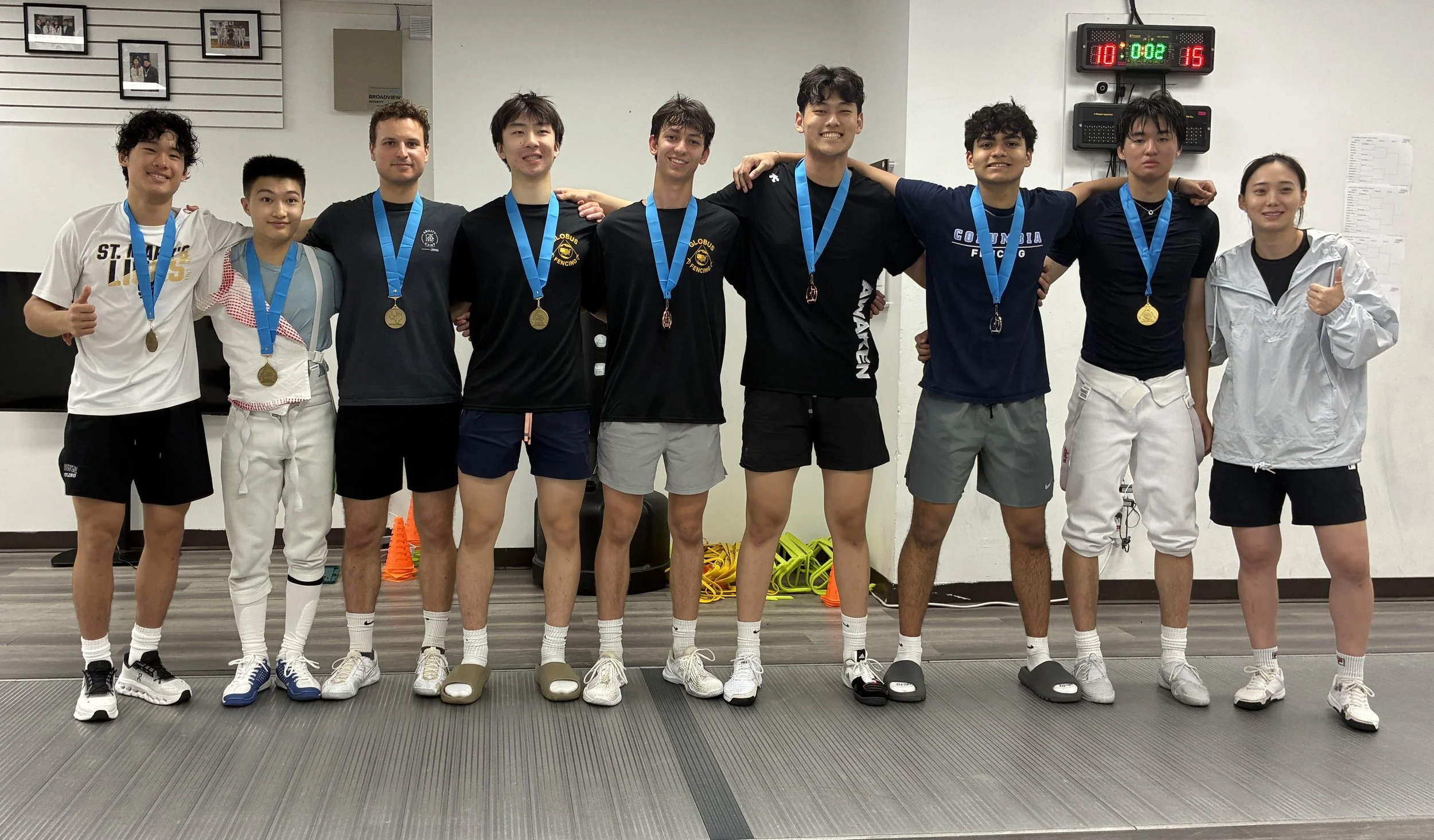 Group of nine young athletes wearing medals, standing in a line with arms around each other, smiling after a competition in an indoor sports facility.