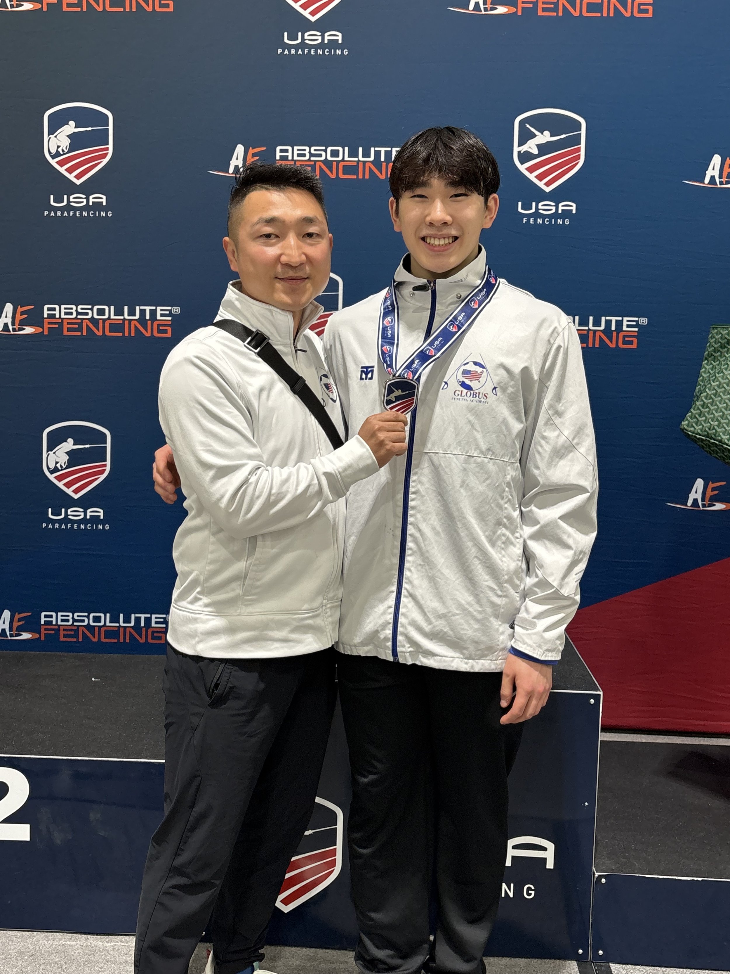 Two men in white jackets standing on a podium at a fencing event, one holding a medal, with a blue backdrop featuring logos for USA Fencing and Absolut Fencing.