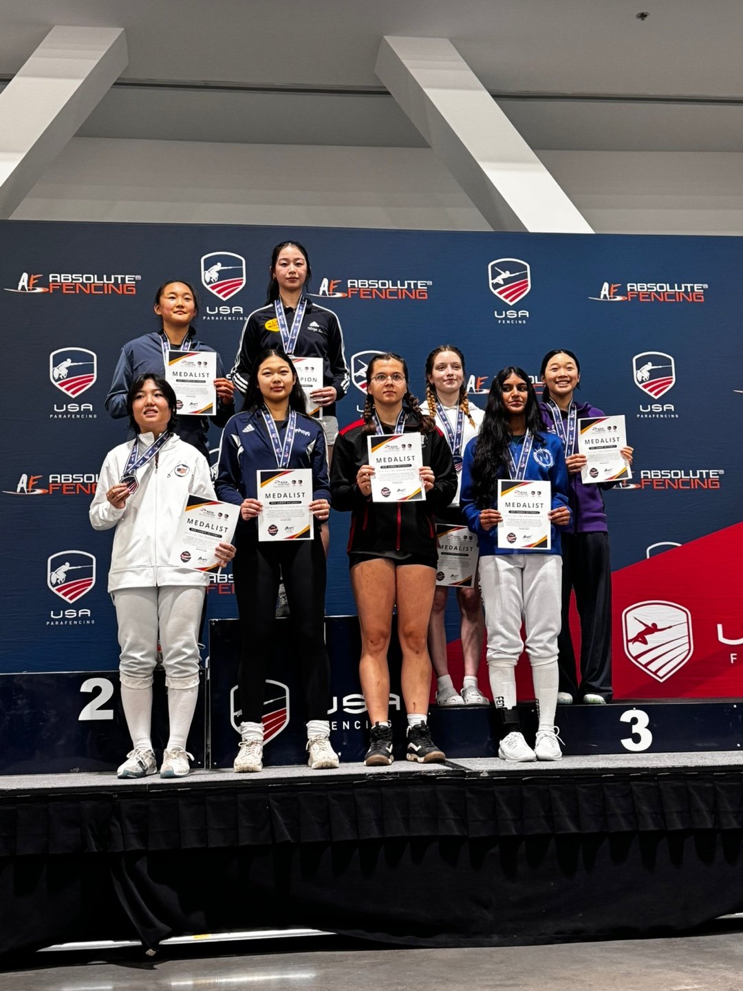 Group of female athletes on a winners' podium holding medals and certificates at a fencing competition.