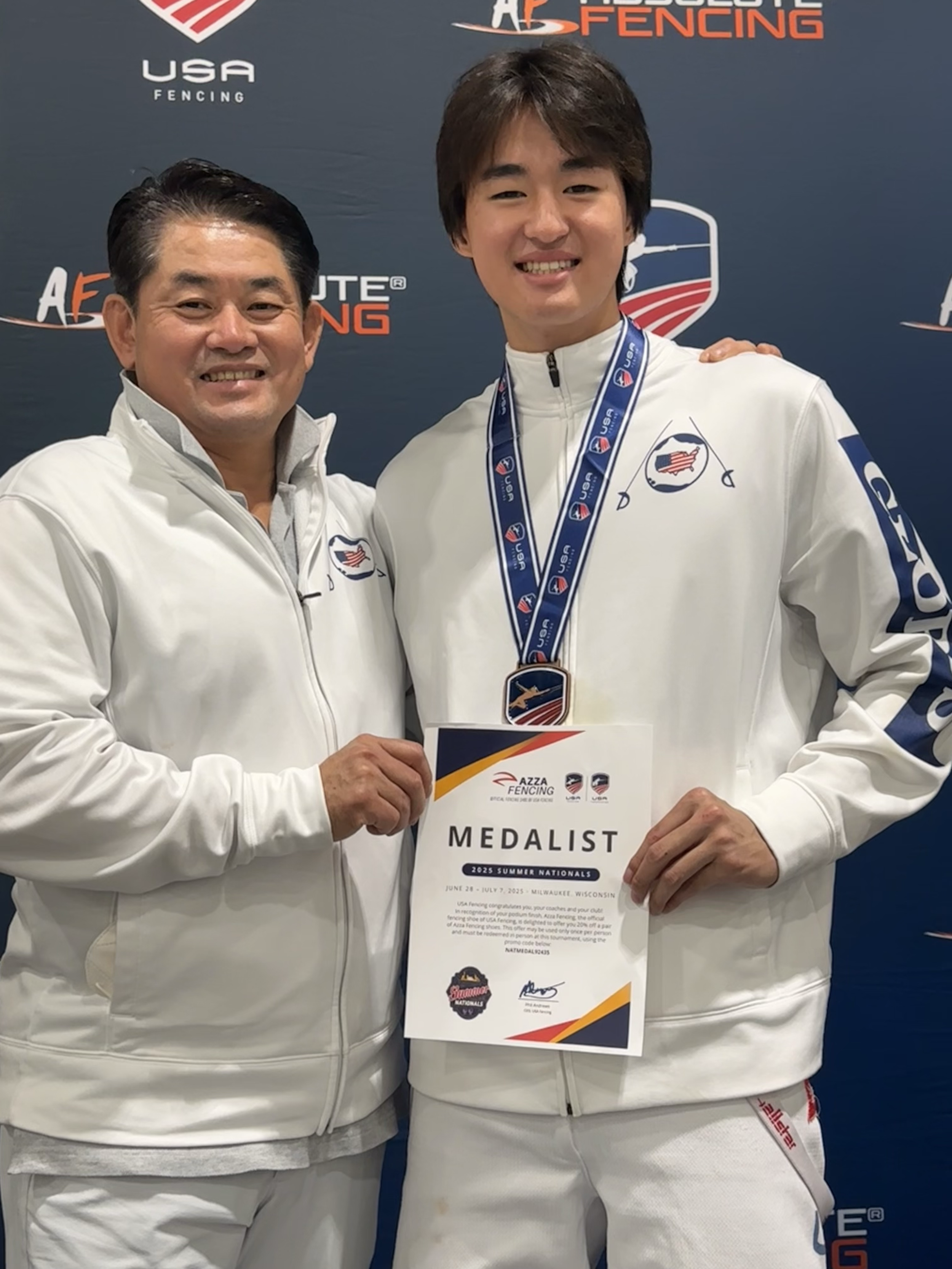 Two people smiling and posing for a photo, one holding a certificate and wearing a medal around his neck, both dressed in white athletic jackets with USA fencing logos, standing in front of a backdrop with USA fencing and logo graphics.