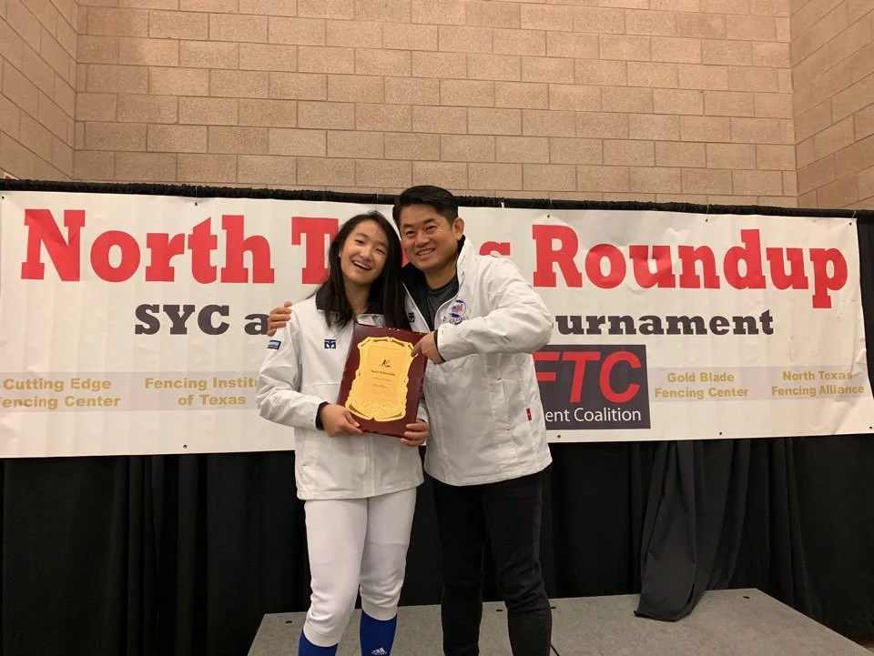 A young woman and a man wearing white fencing jackets are smiling and holding a plaque, standing on a stage in front of a banner that reads 'North Texas Roundup, SYC and Tournament'.